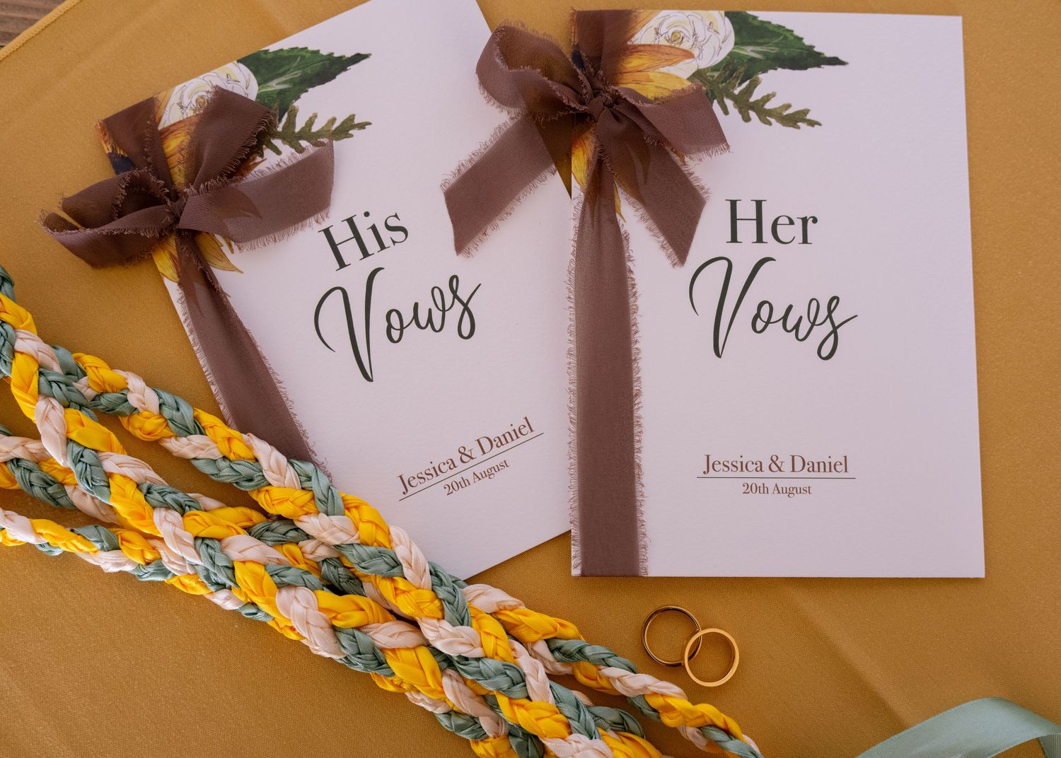 His and Her Vows booklets with brown ribbon bows beside braided handfasting cord and gold wedding rings on yellow fabric.
