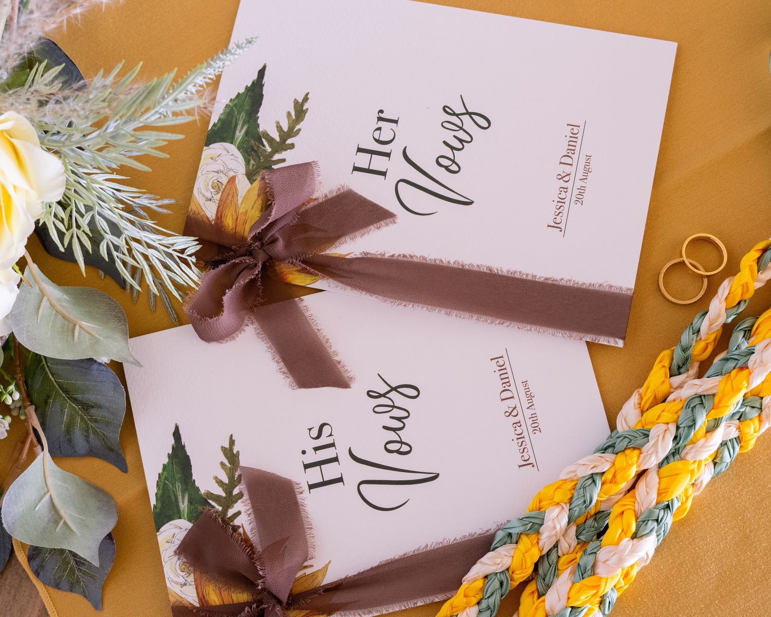Elegant wedding vow books with brown ribbon, floral decor, gold rings, and braided cord on golden background.