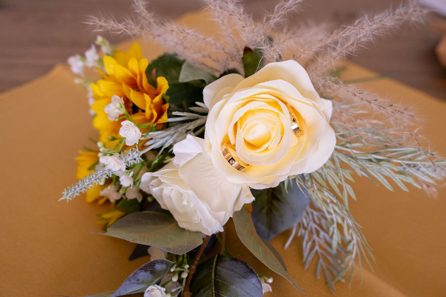 A elegant floral arrangement featuring cream roses, yellow sunflowers, and pampas grass on a golden background.