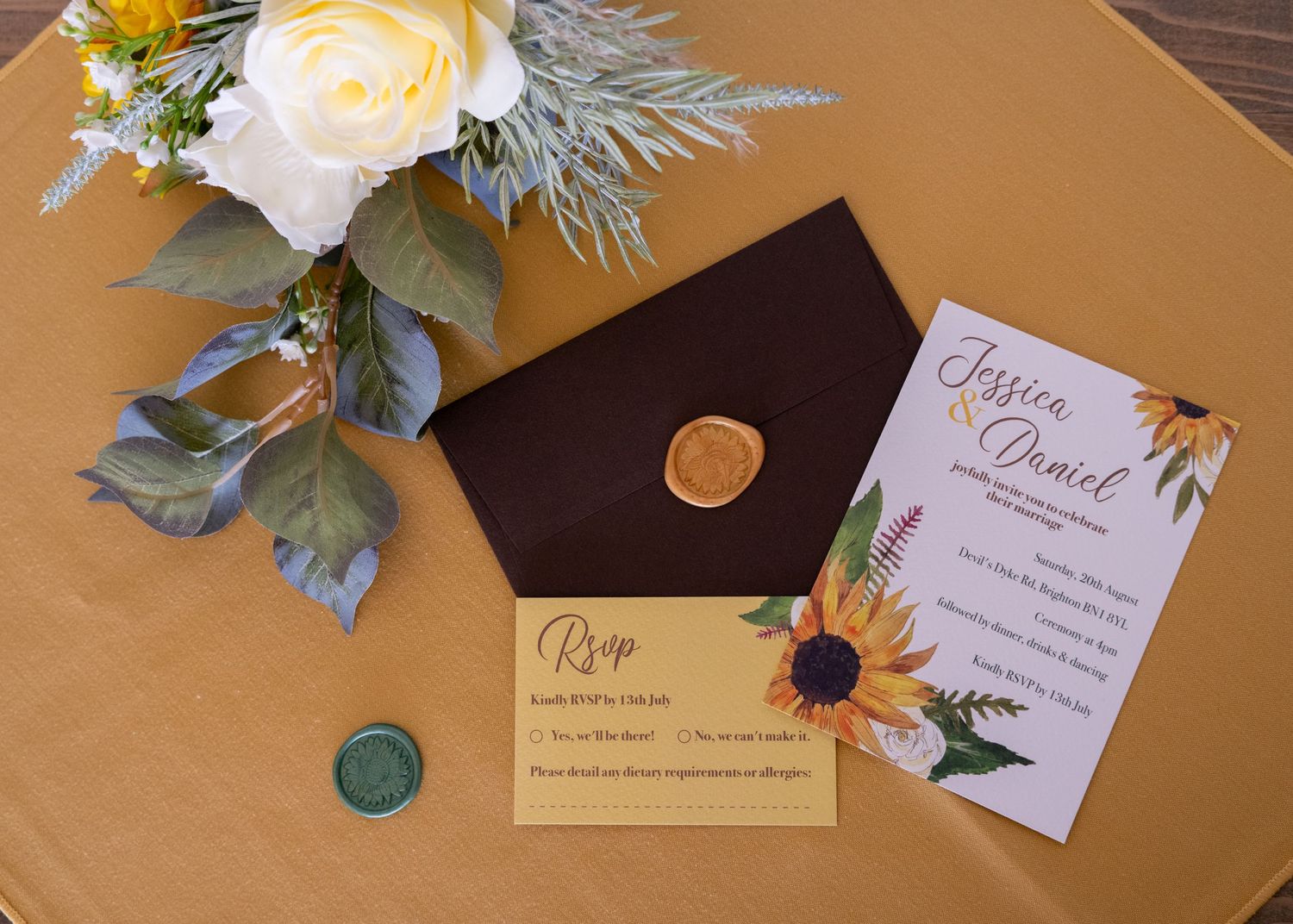 Elegant wedding invitation suite with sunflower design, dark envelope with wax seal, RSVP card, and floral bouquet on gold tablecloth.