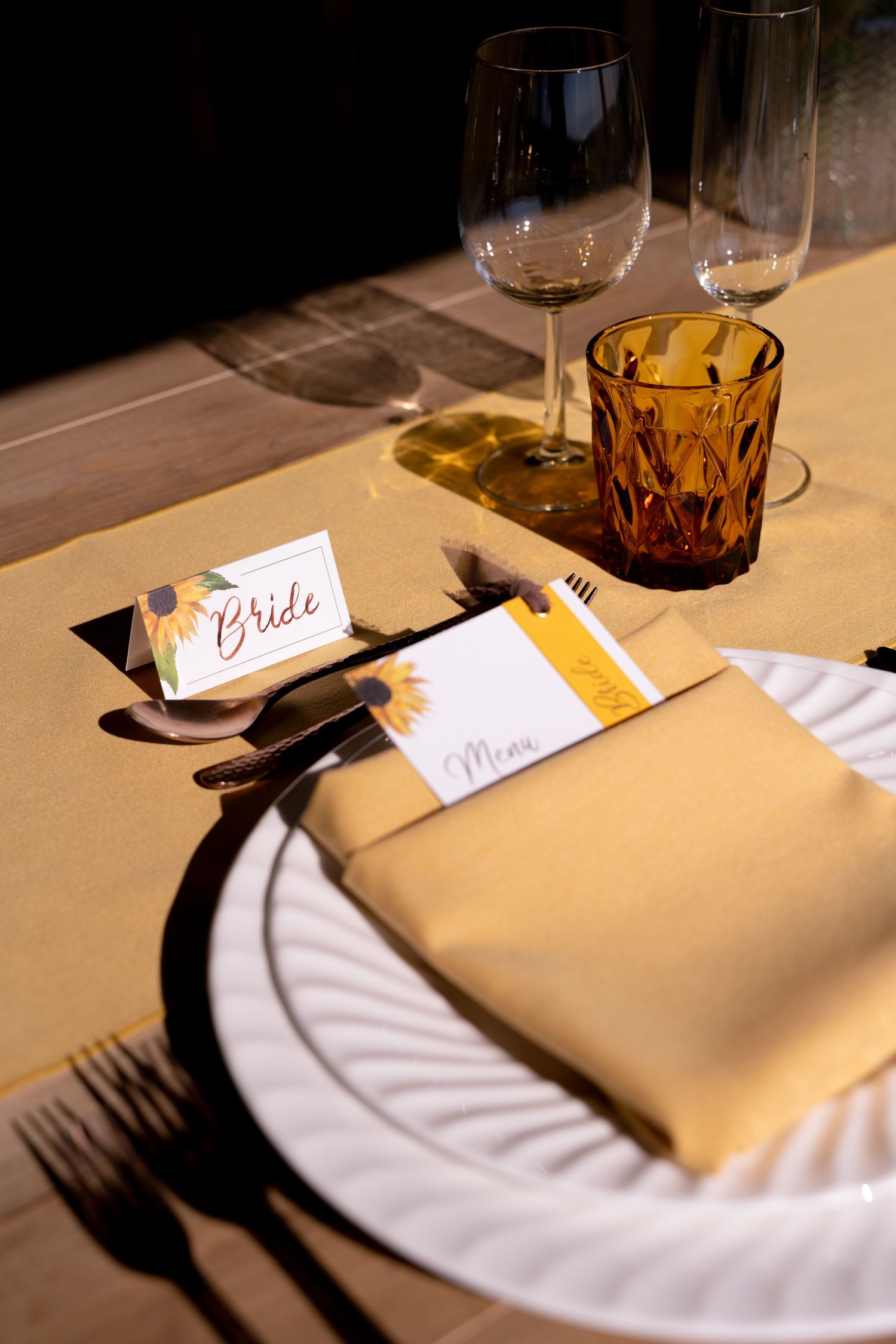 Elegant wedding table setting with sunflower-themed bride place card, yellow napkin, white plates, amber glass, and wine glasses.