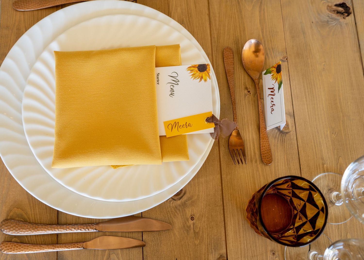 Elegant sunflower-themed table setting with gold napkin, copper cutlery, place cards, and decorative candle holder.