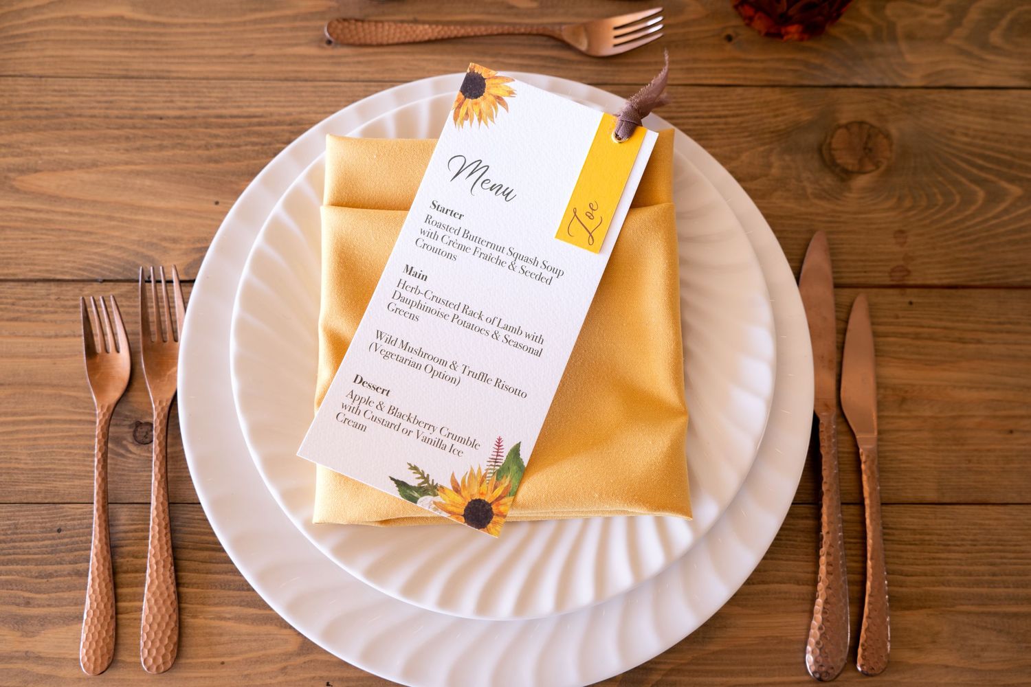 Elegant fall wedding place setting with gold cutlery, white plates, yellow napkin, and sunflower menu card on wood table.