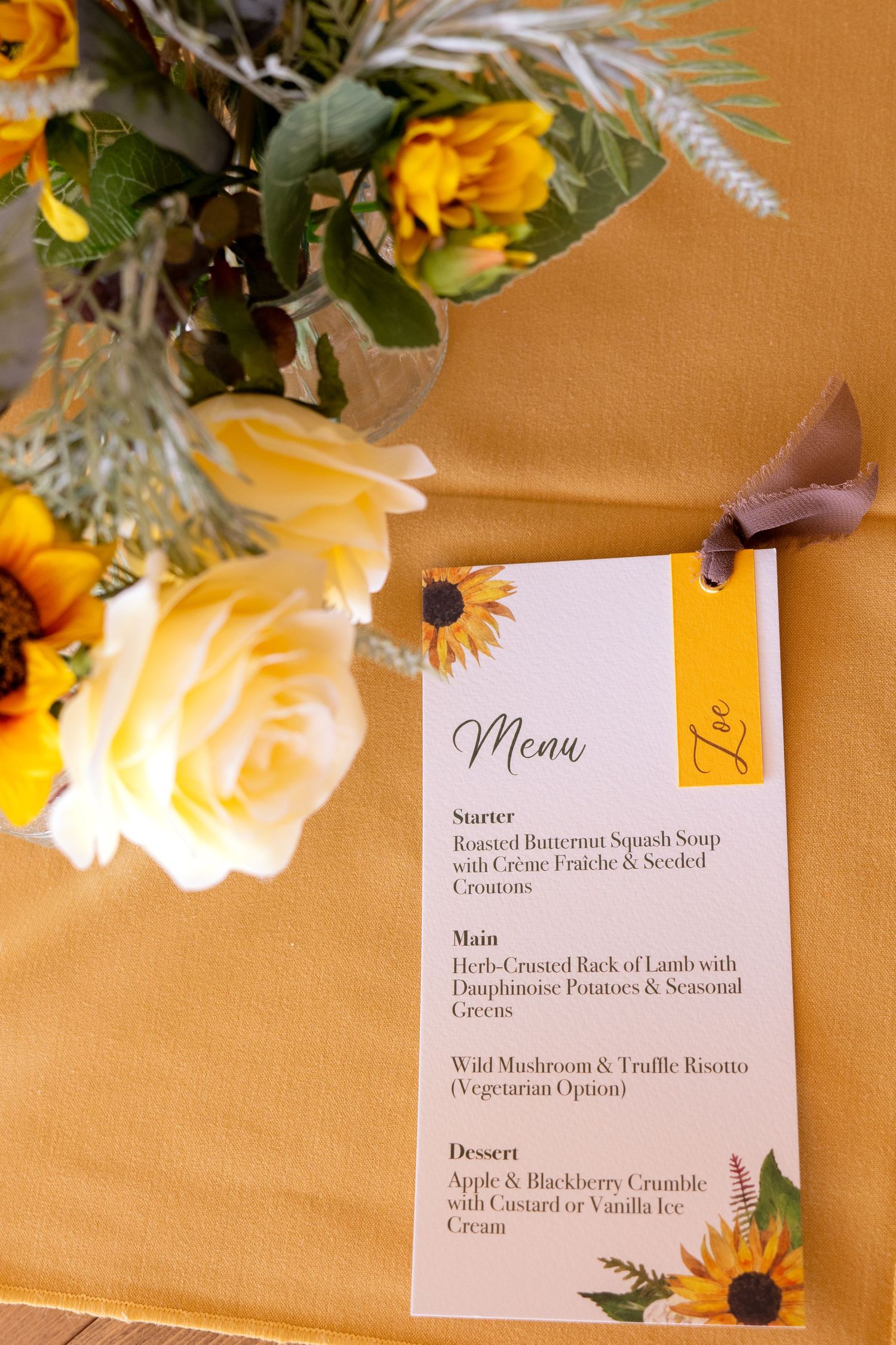 Wedding menu card with sunflower design beside yellow roses and sunflowers on a golden tablecloth.