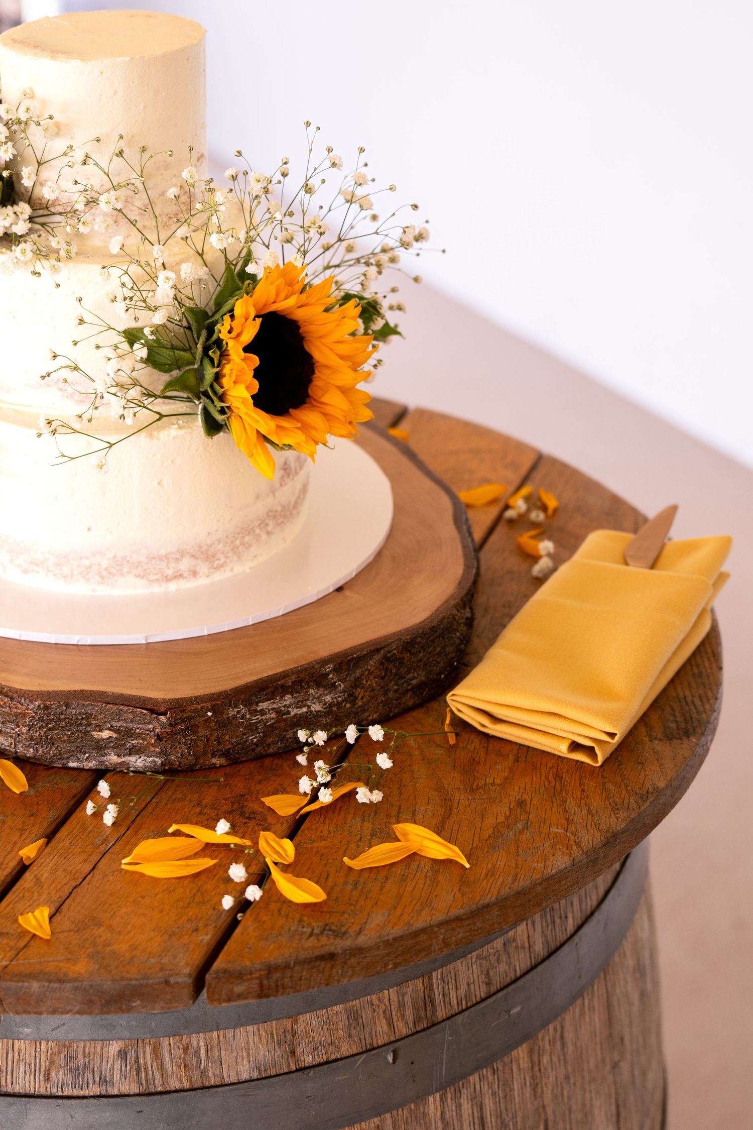 White two-tier wedding cake with sunflower topper on rustic wood slice atop a barrel, with yellow napkin and petals.