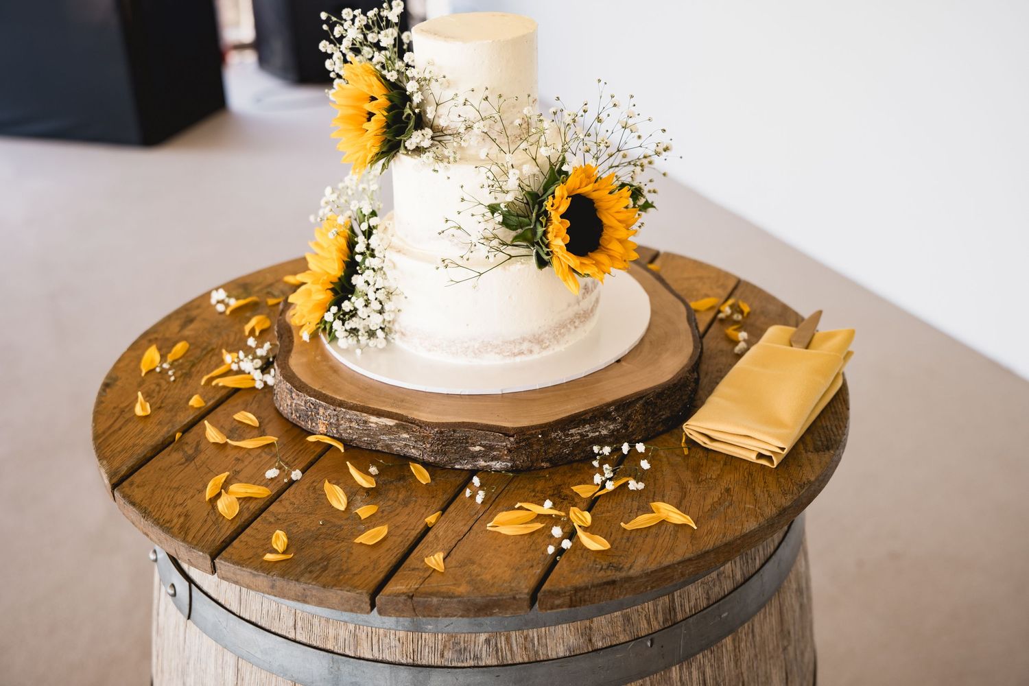 White wedding cake with sunflowers and baby's breath on rustic wood barrel stand with scattered yellow petals.