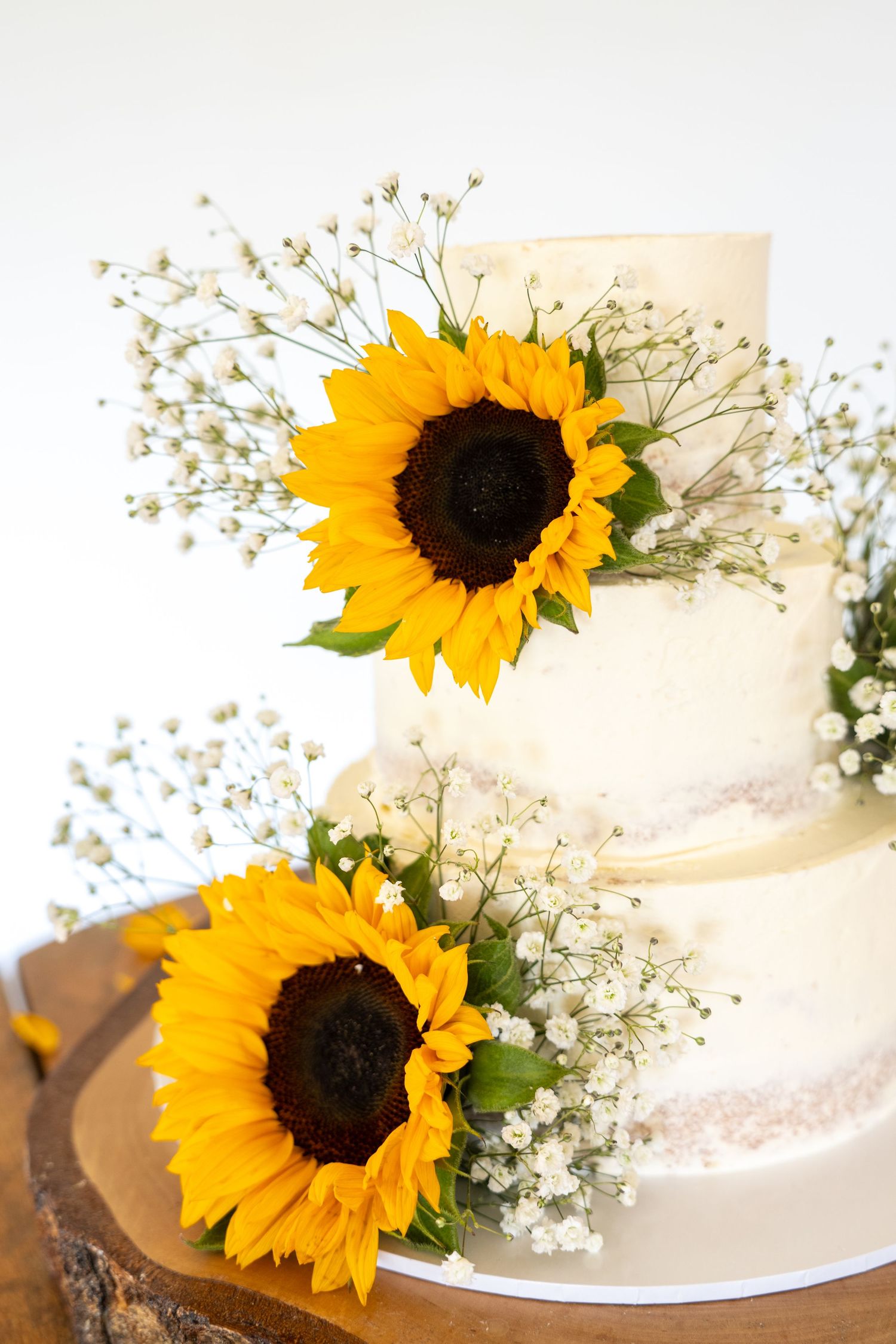 Three-tier white wedding cake decorated with bright yellow sunflowers and baby's breath on a rustic wood slice.