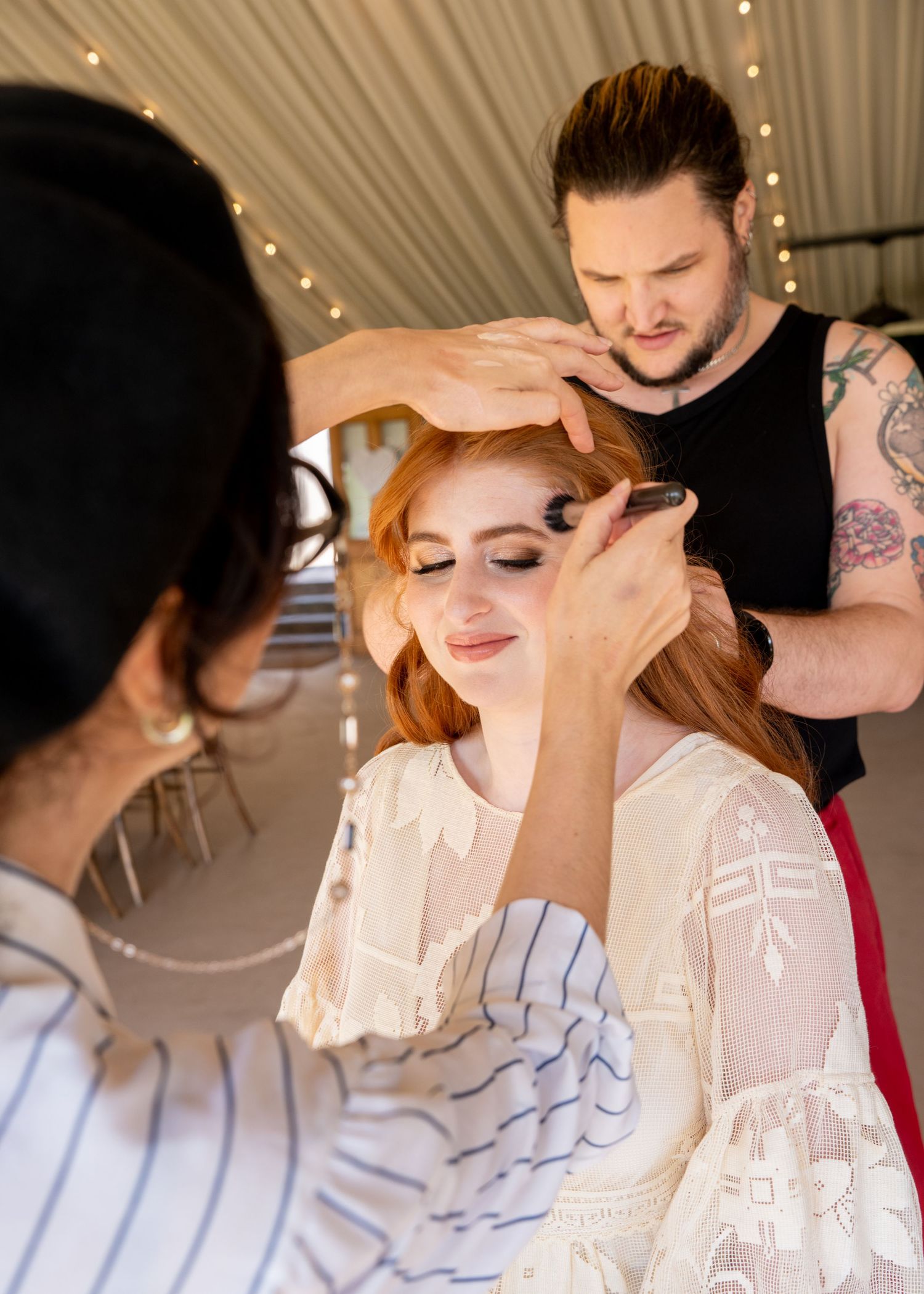 A redheaded woman gets hair and makeup done by two stylists in a bright tent with fairy lights.