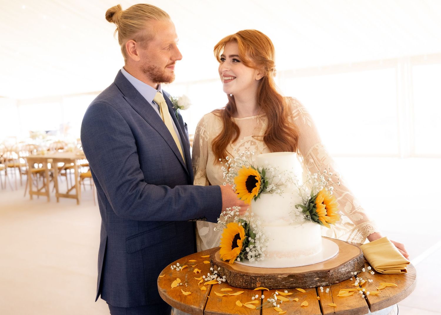 Bride and groom cutting white wedding cake decorated with sunflowers on rustic wood stand in elegant tent venue.