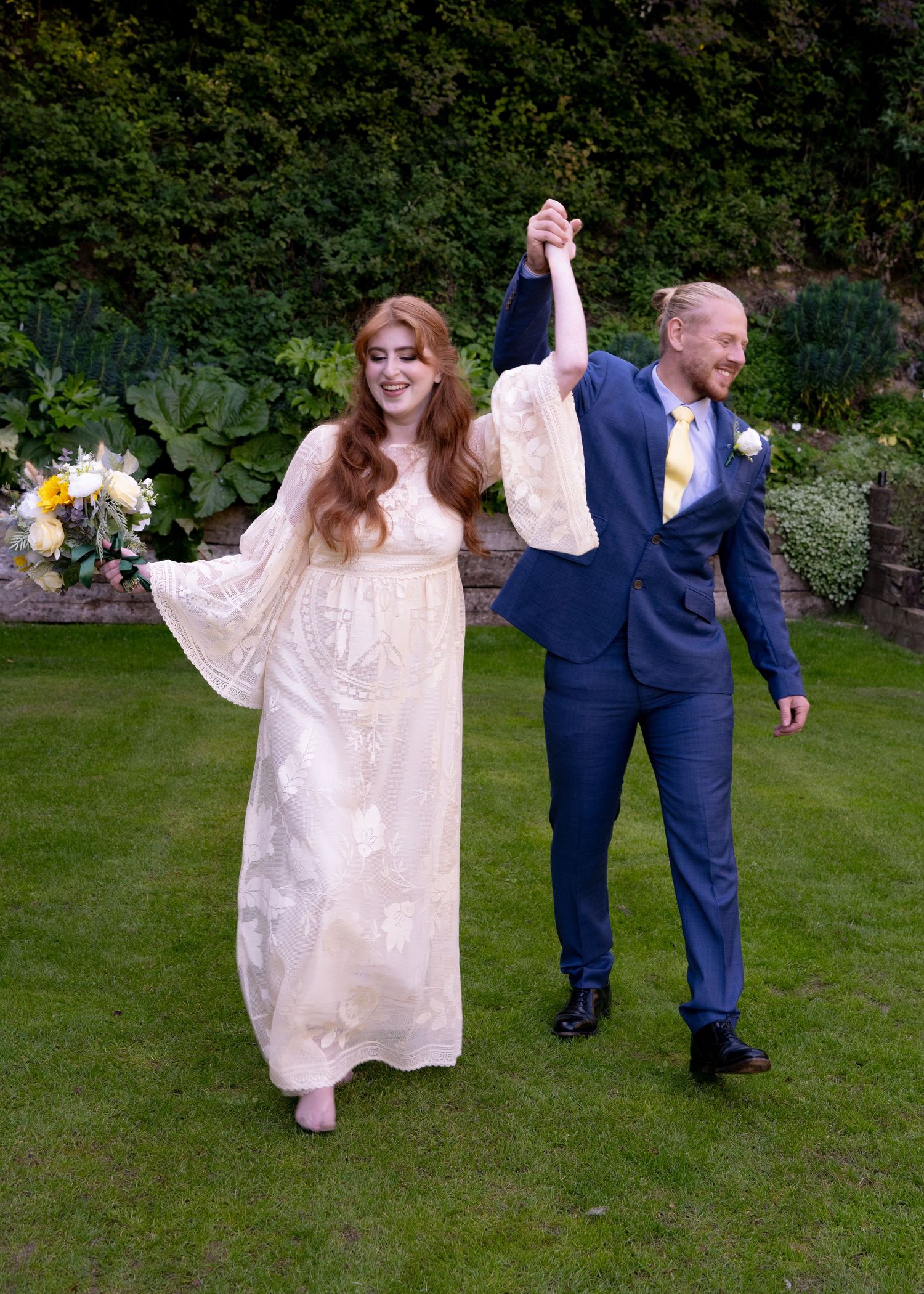 Bride and groom celebrate with raised hands on garden lawn, bride holding white and yellow bouquet.