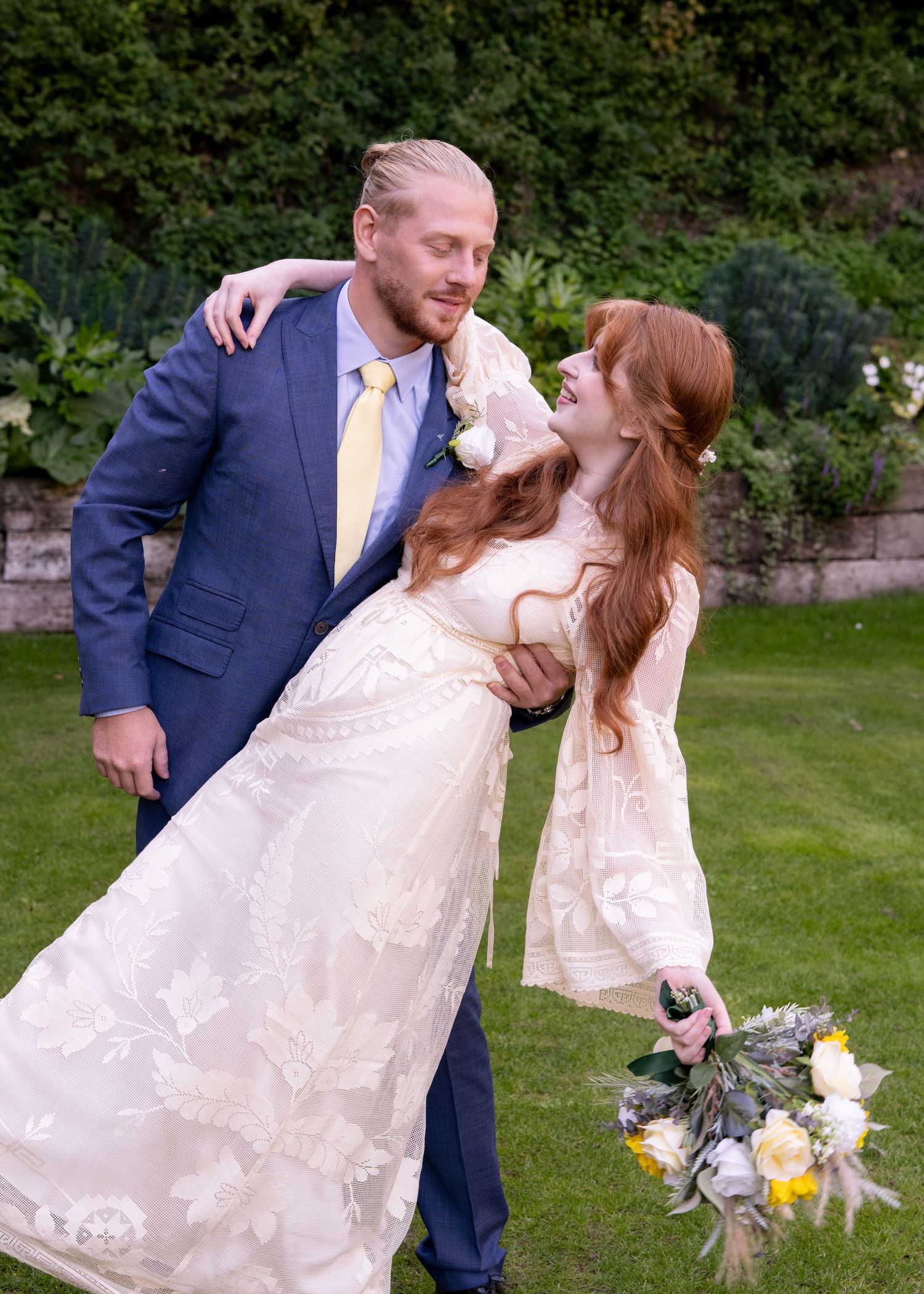 Bride in lace gown holding yellow bouquet gazes at groom in blue suit on lush garden lawn.