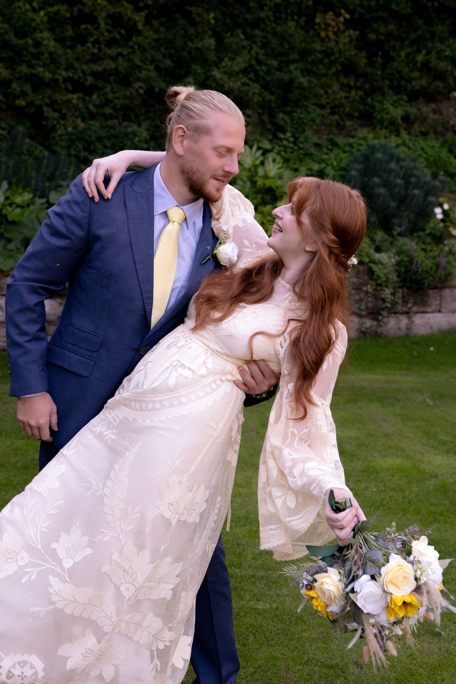 Bride in lace gown and groom in blue suit embrace lovingly in a lush green garden.