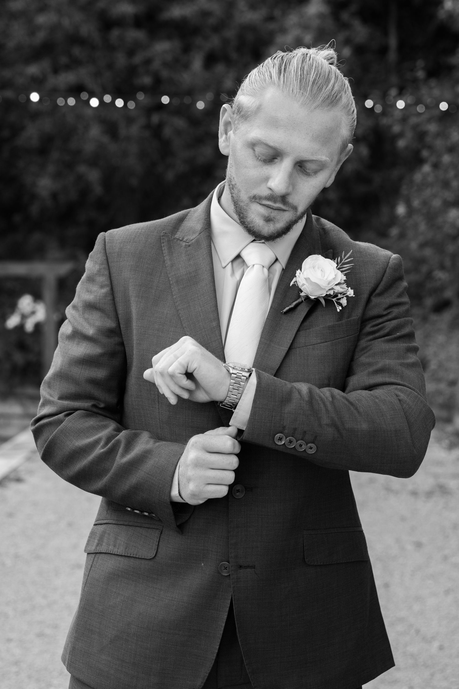 Groom in gray suit adjusting watch, wearing boutonniere, black and white wedding photo outdoors.