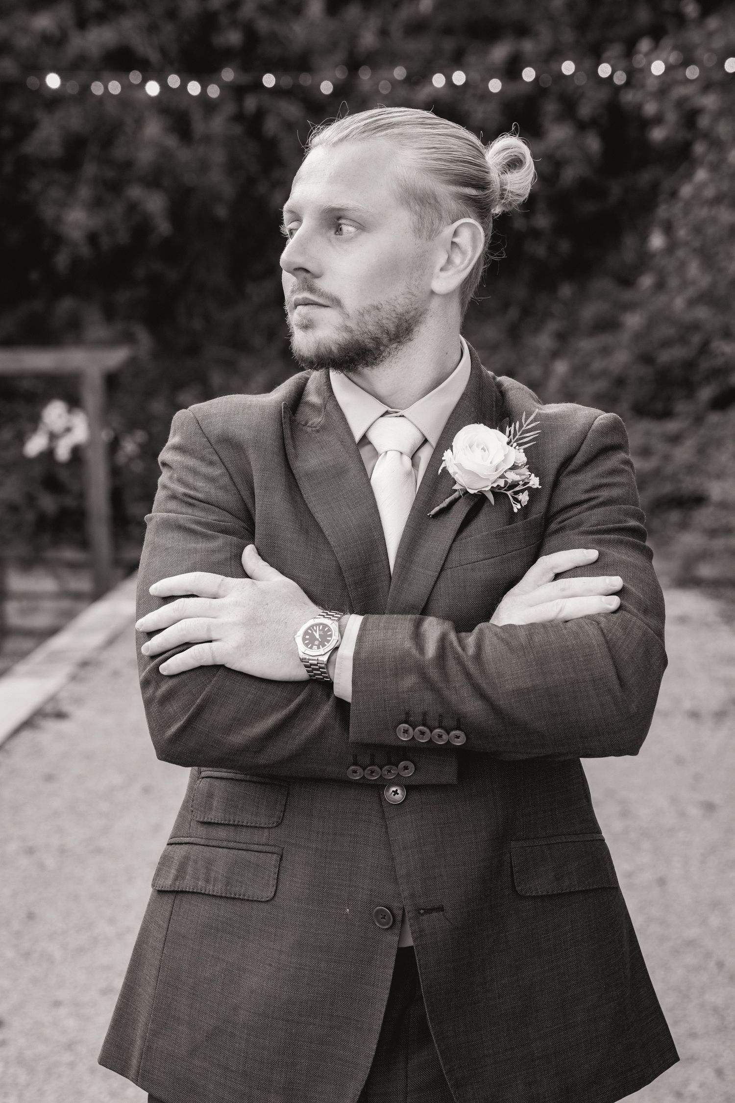 Groom in a suit with boutonniere and man bun, arms crossed, posing outdoors at a wedding venue.