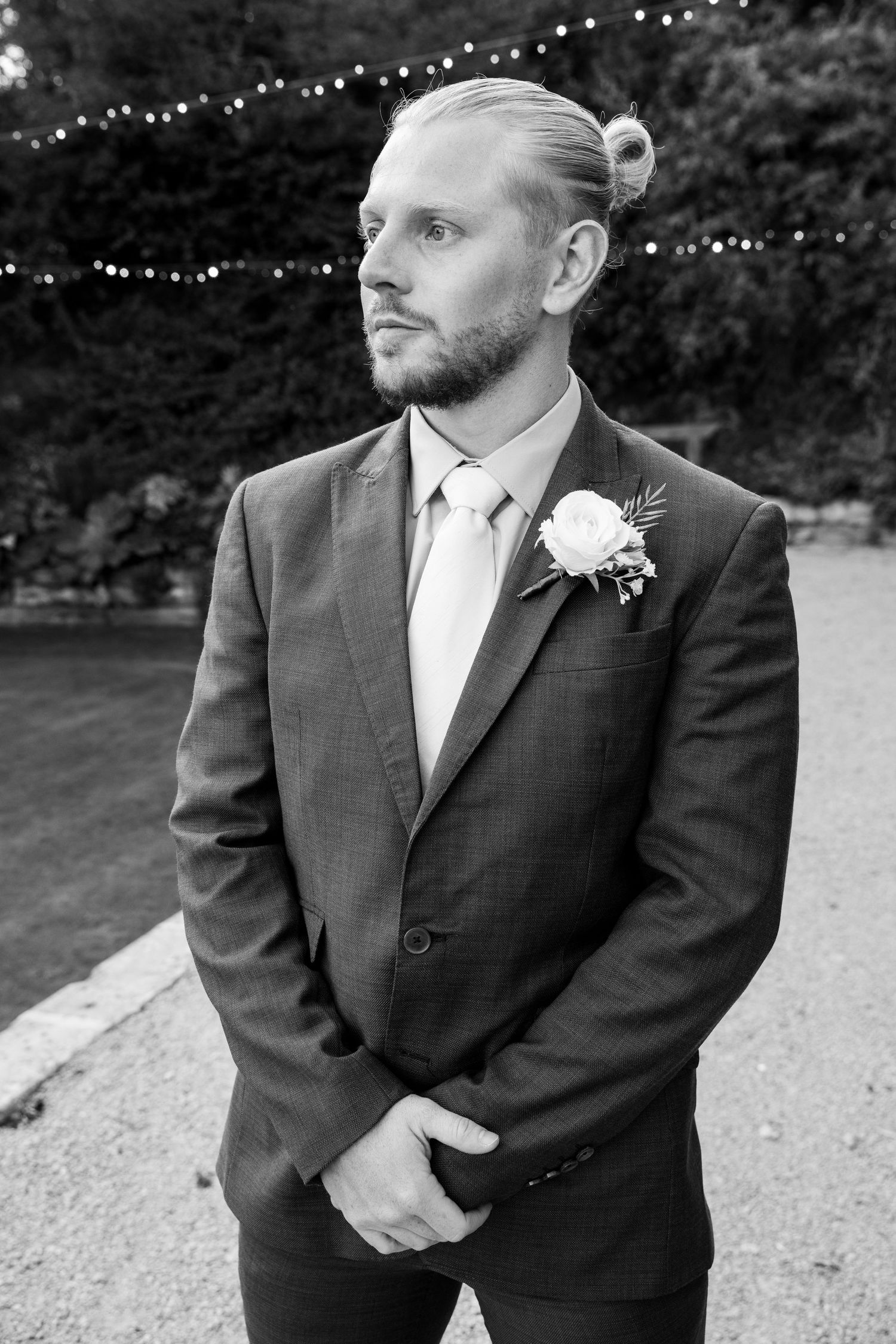 Black and white portrait of groom in suit with rose boutonniere and hair bun outdoors.