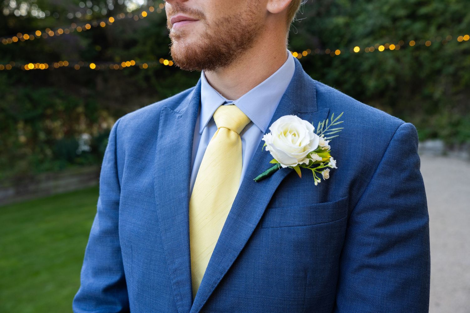 Man in blue suit with yellow tie and white rose boutonniere at outdoor wedding ceremony.