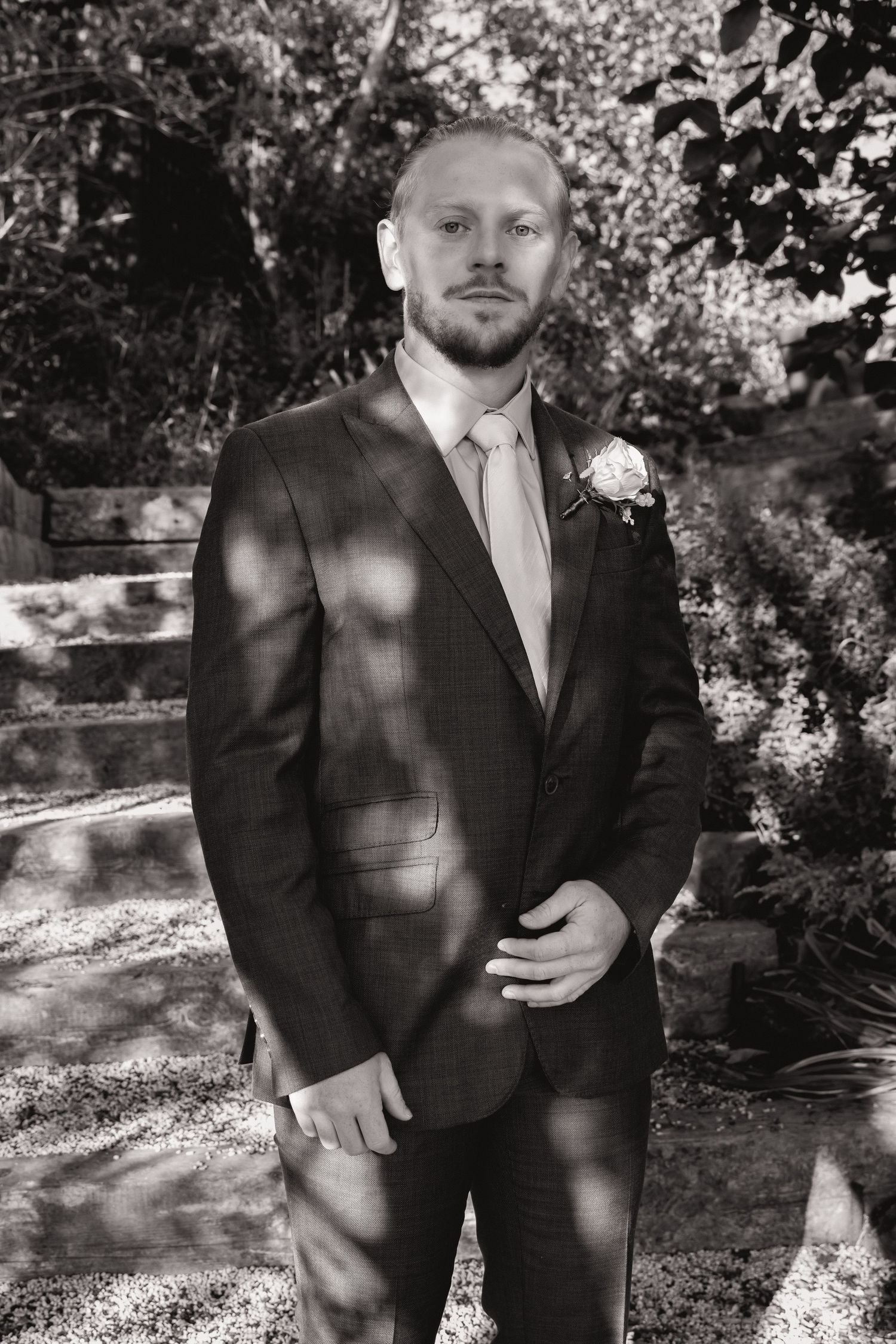 Black and white portrait of a groom in a suit with boutonniere standing outdoors among trees.