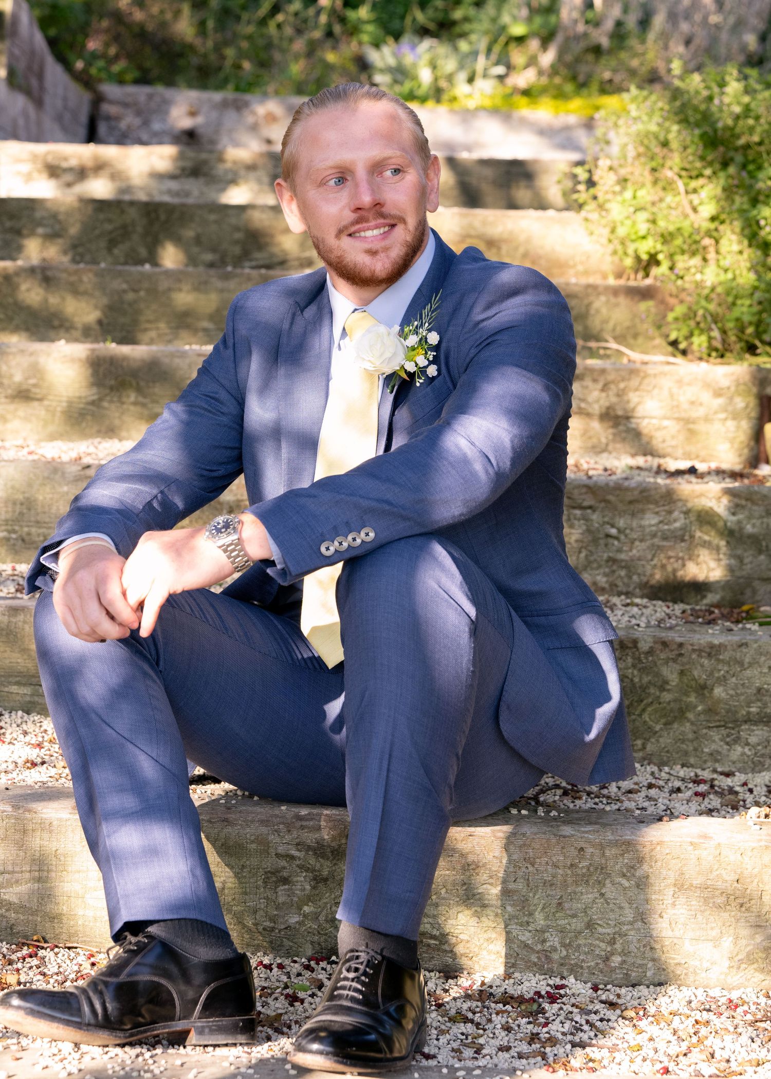 Smiling groom in blue suit with boutonniere sitting on stone steps outdoors.