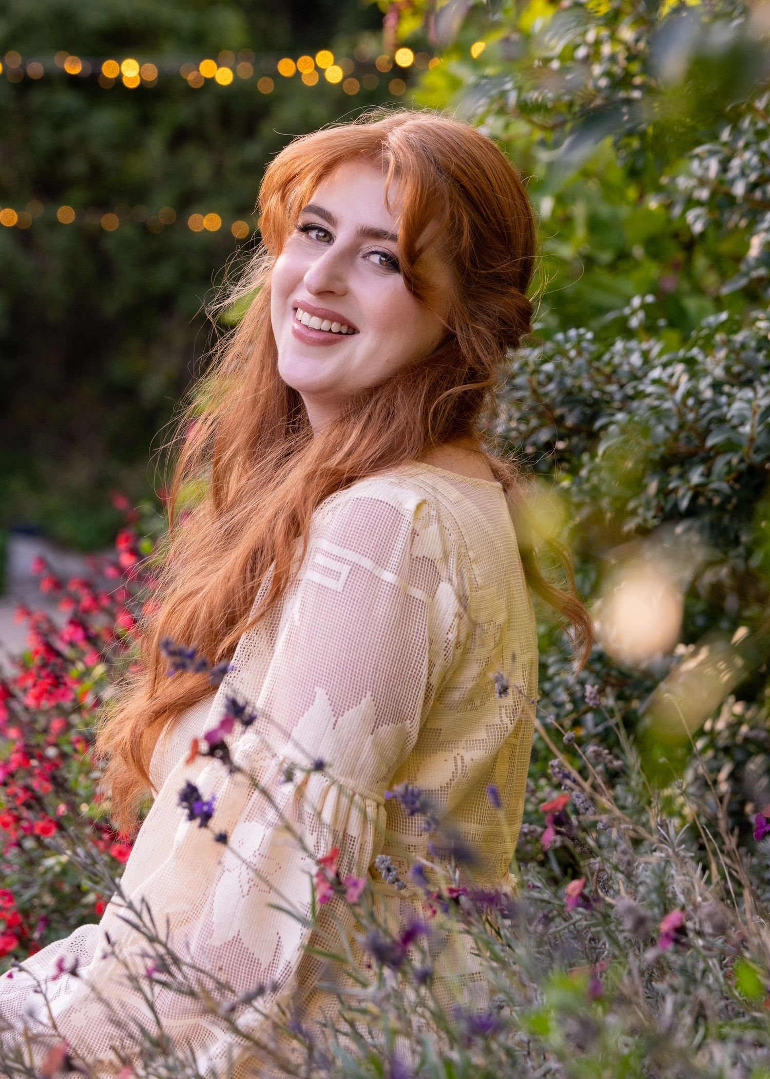Smiling redhead woman in cream lace dress surrounded by colorful garden flowers and bokeh lights.