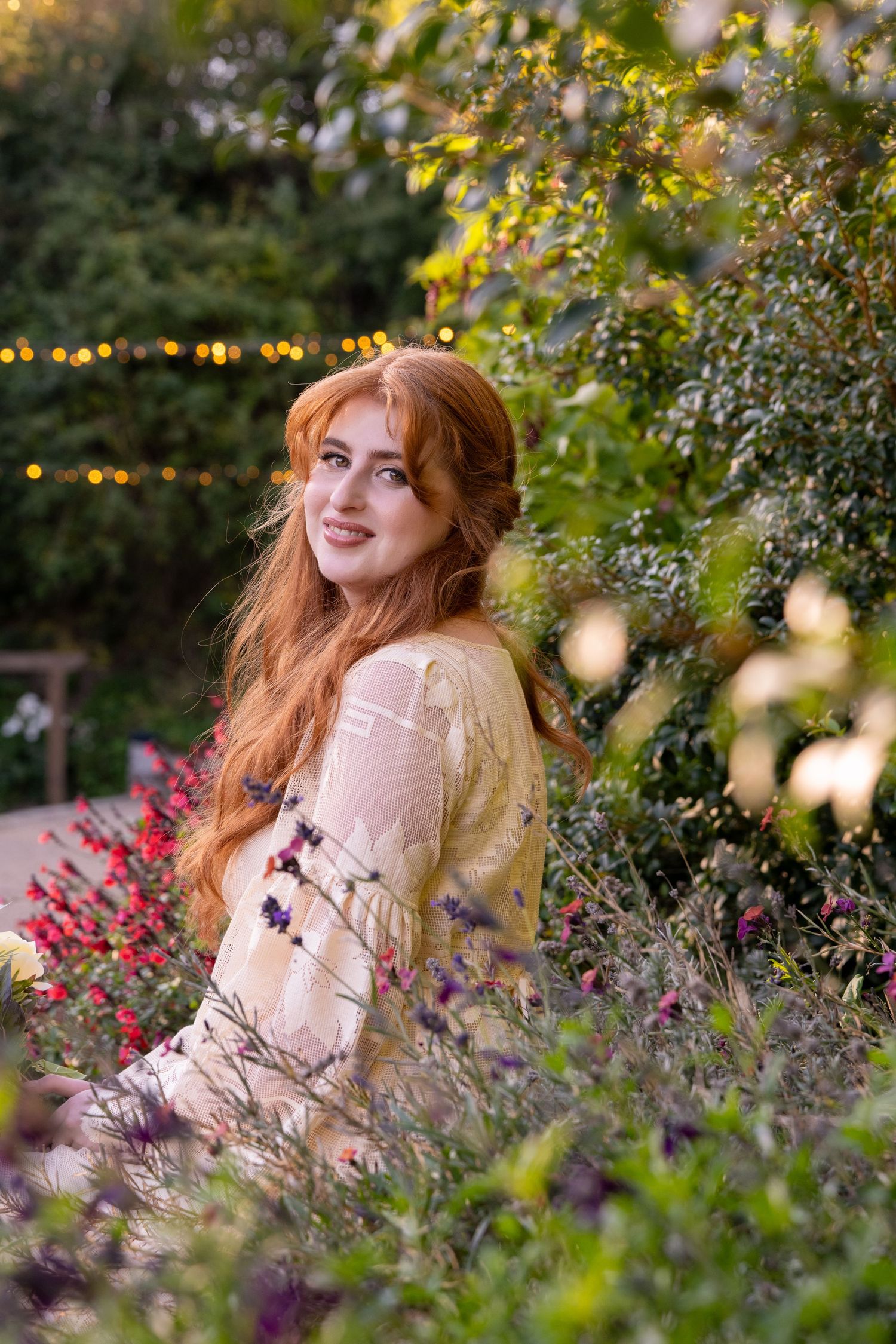 Young woman with long red hair smiling among colorful garden flowers at dusk.