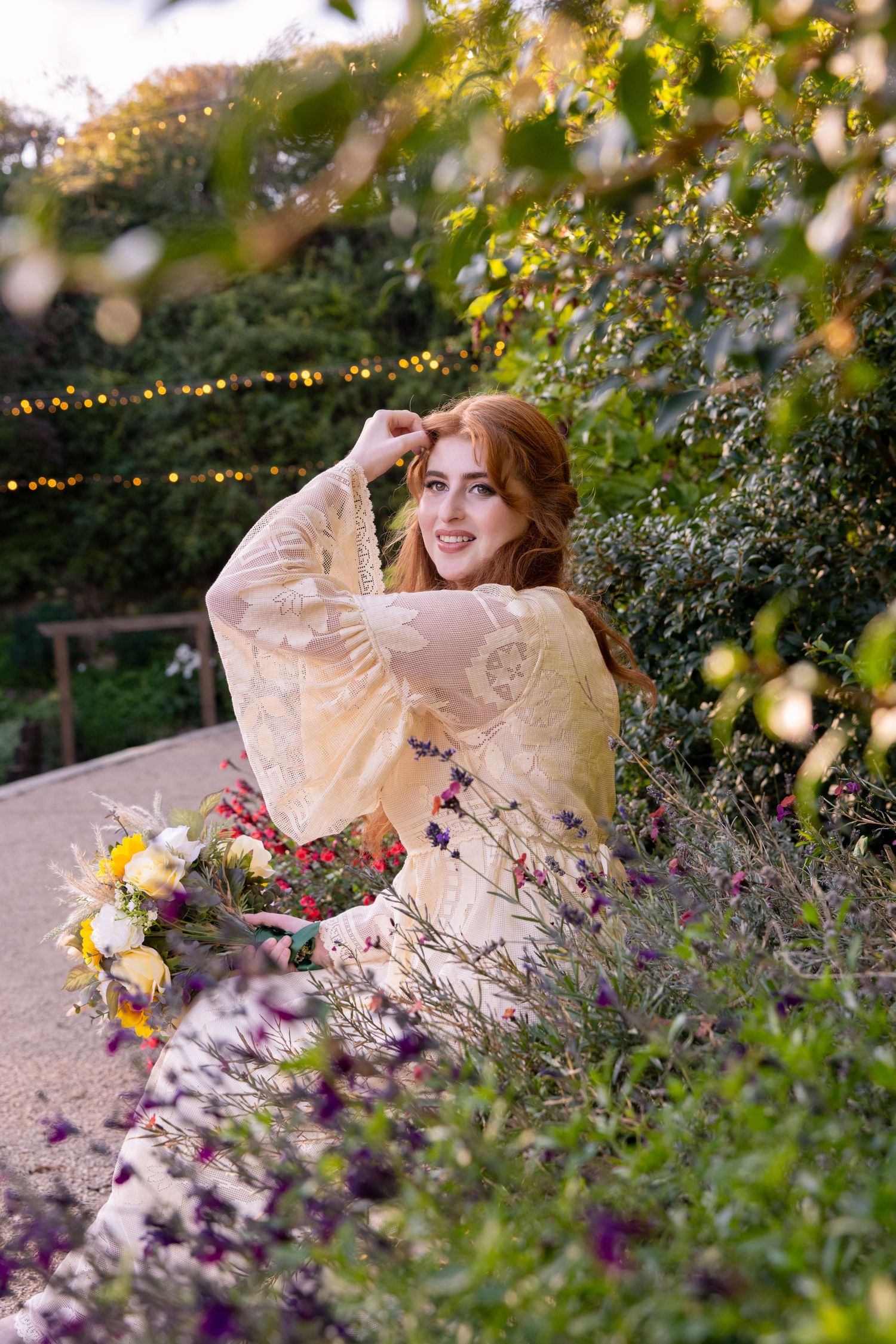 A redhead woman in a yellow lace dress sits among colorful garden flowers with string lights behind her.