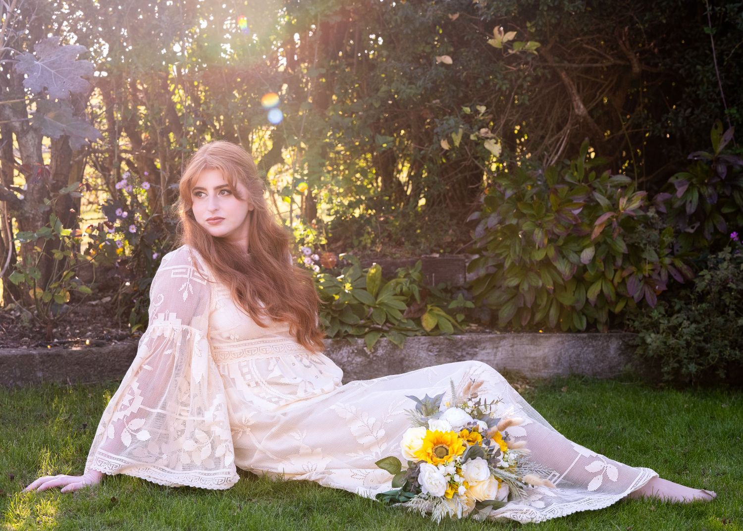 Woman in lace boho wedding dress sitting on grass with sunflowers, bathed in golden sunlight among green foliage.