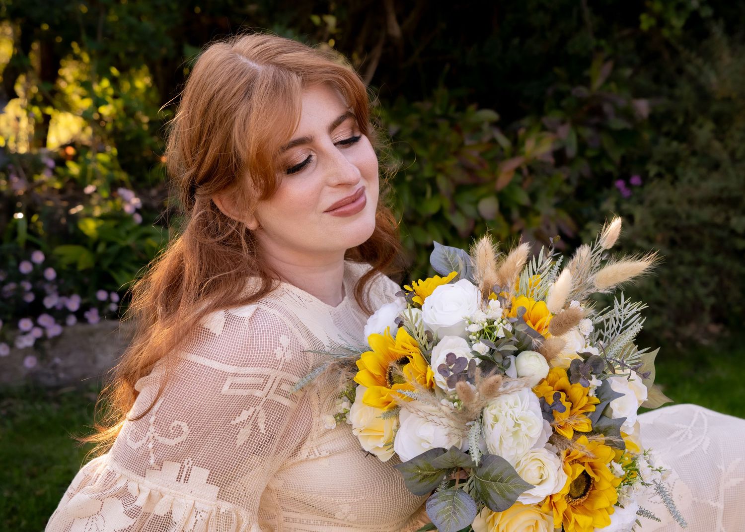 Bride with red hair holding a bouquet of sunflowers and white roses, wearing a lace dress in a garden setting.