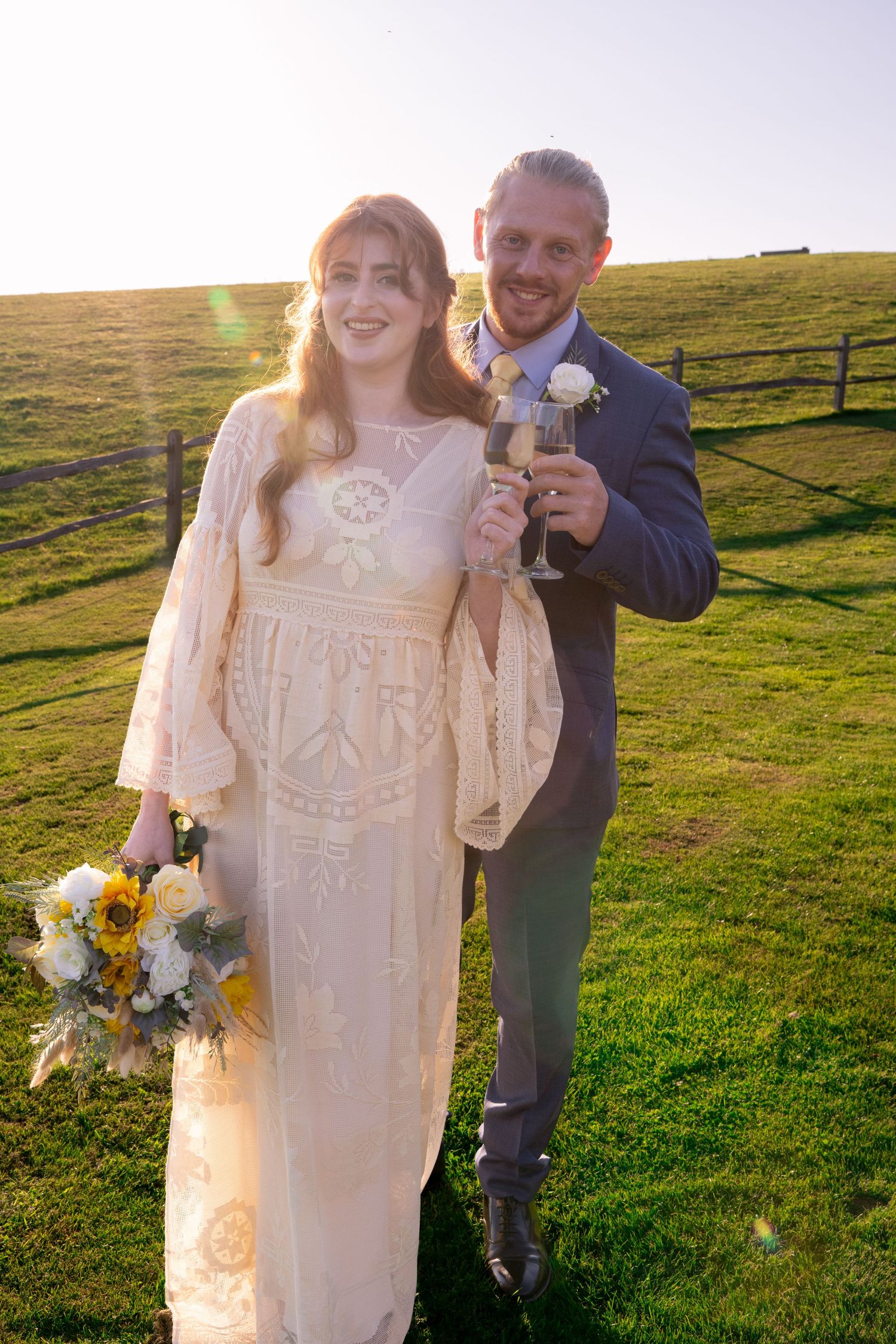 Bride and groom toasting with champagne in sunlit green field on wedding day.