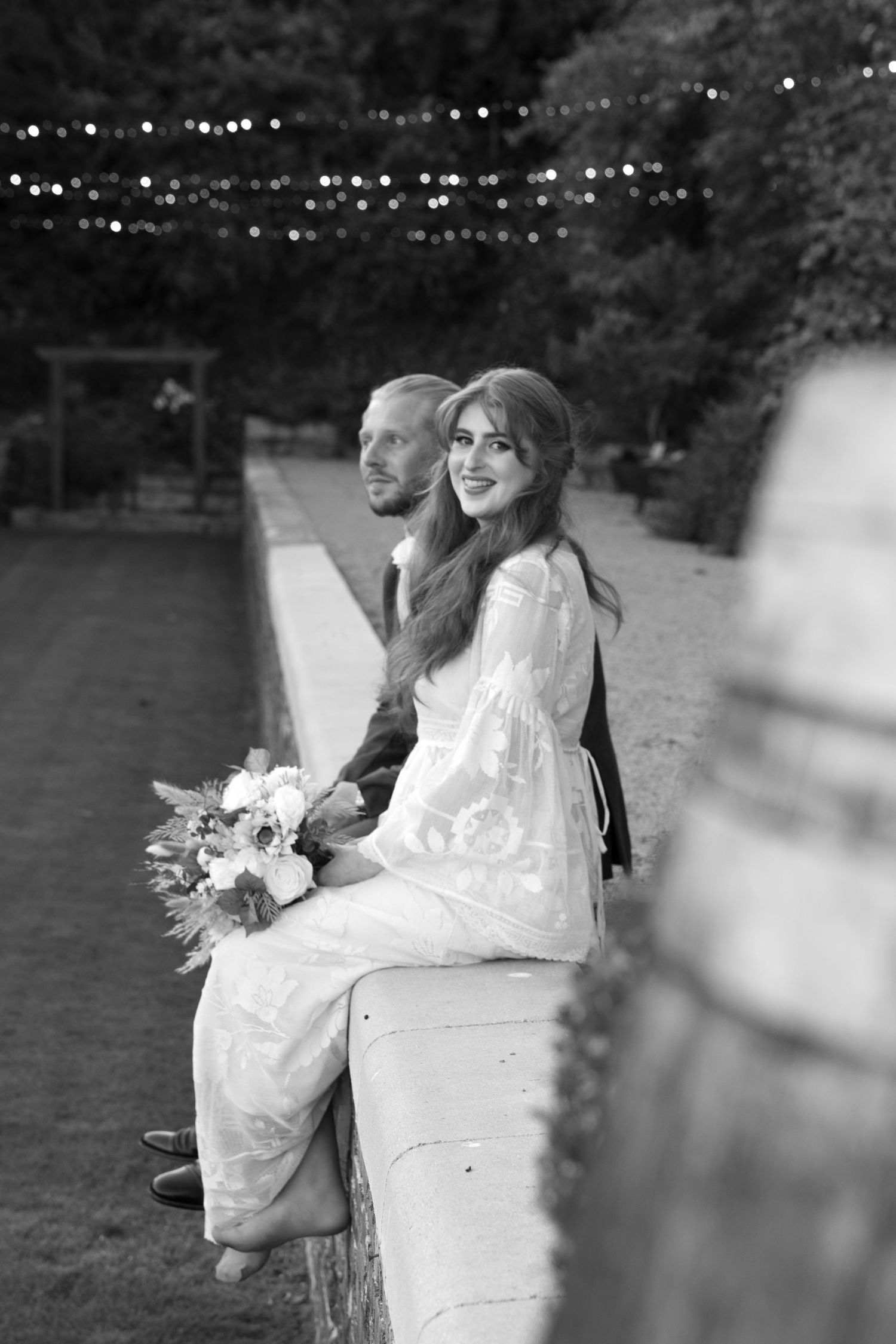Bride in white dress holding bouquet sits with groom outdoors at wedding, string lights in background.