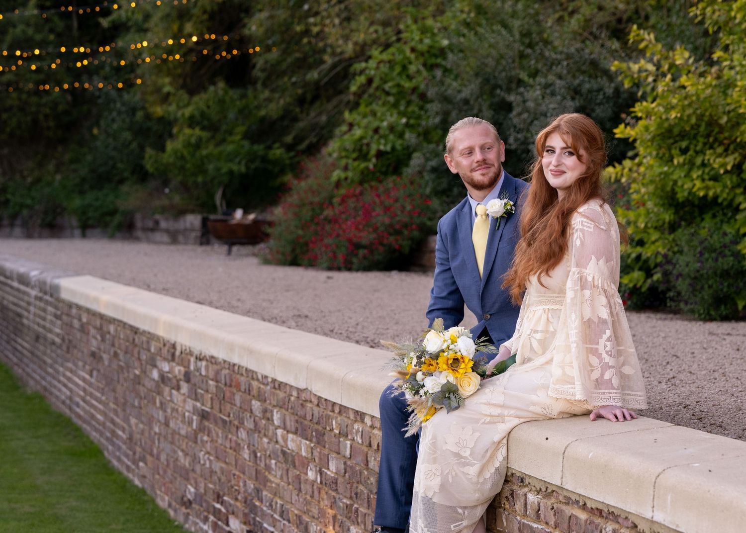 Smiling bride and groom sitting on a stone wall outdoors at their wedding with yellow bouquet.