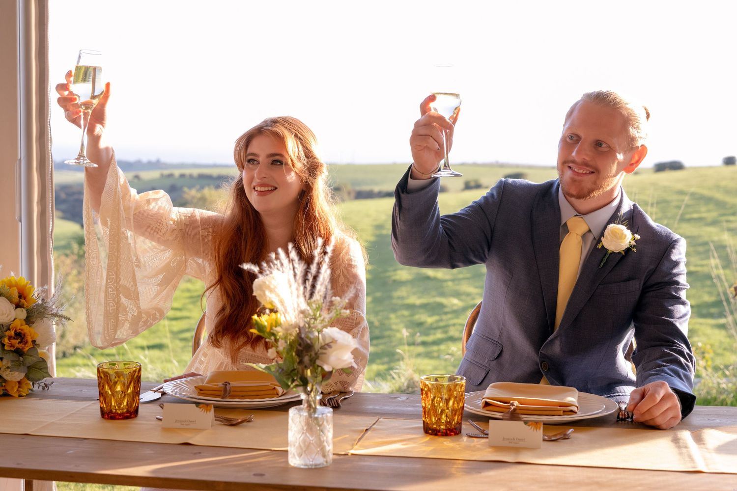 Bride and groom toasting with champagne glasses at rustic wedding table with floral centerpiece and scenic countryside view.