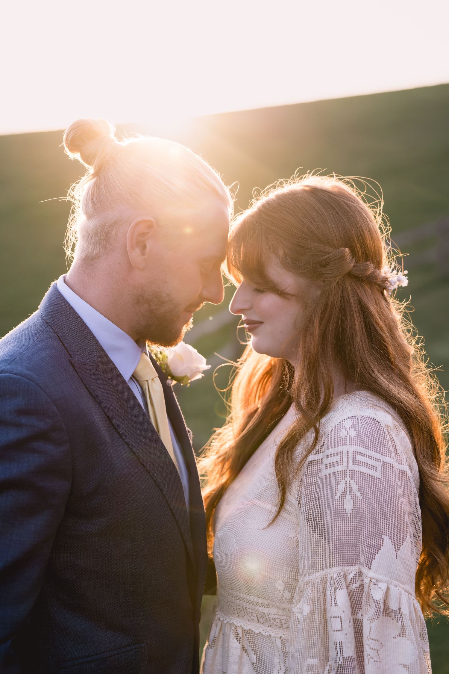 Bride and groom touching foreheads in golden sunset light, she in lace dress, he in navy suit.