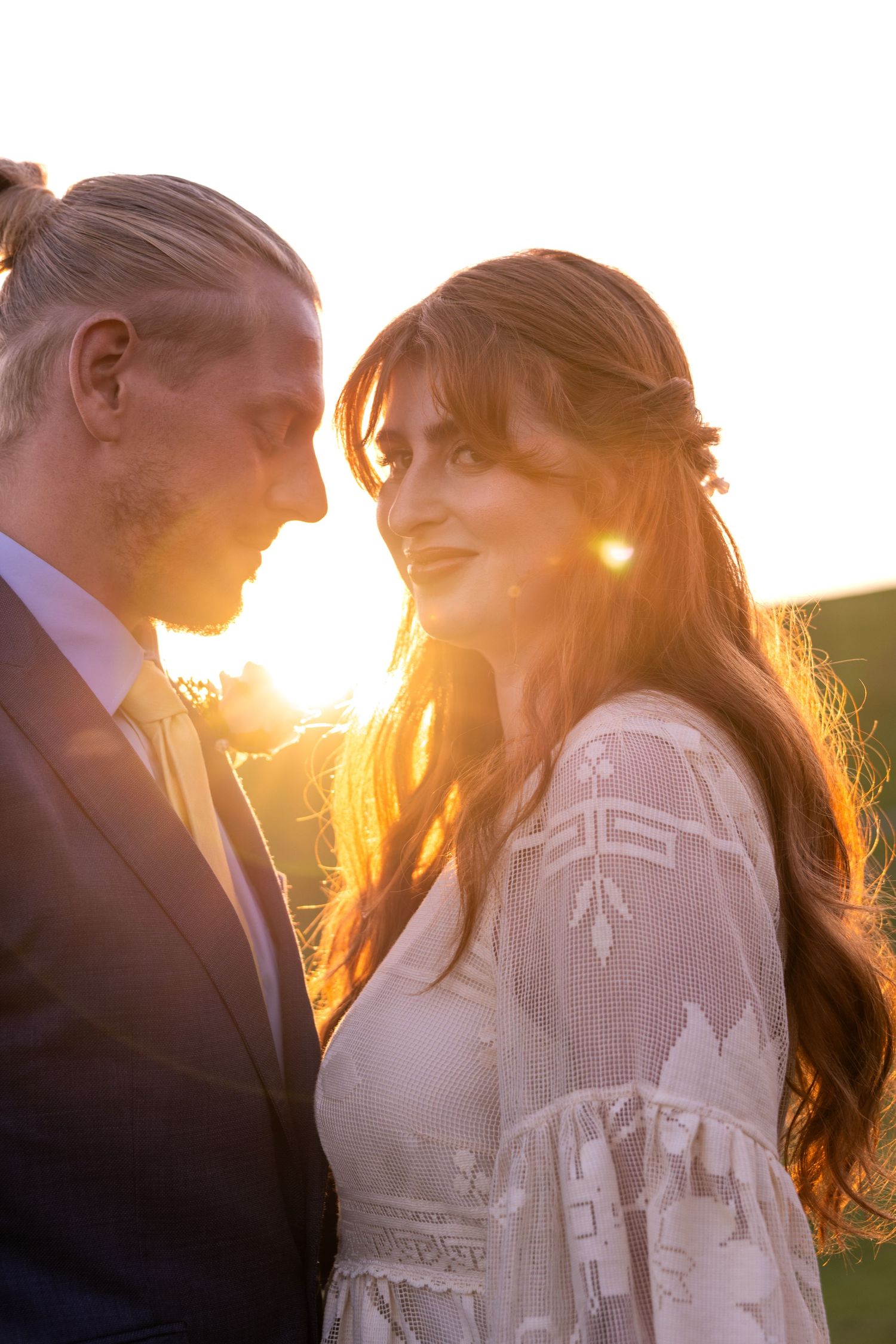 Bride and groom share a romantic golden hour moment with warm sunset light glowing behind them.