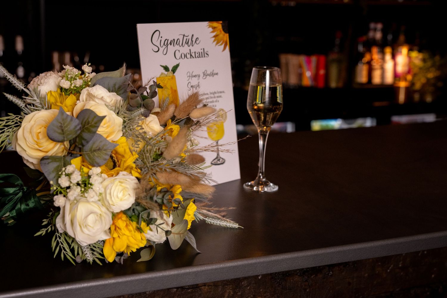 White and yellow wedding bouquet beside signature cocktails menu sign and champagne glass at bar.