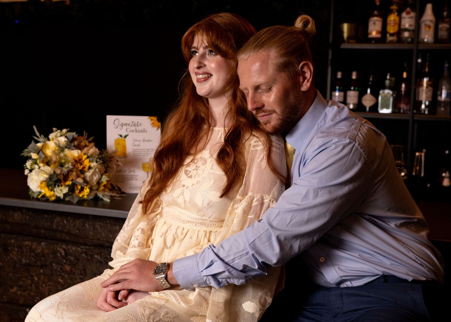 Couple in wedding attire sitting closely together at a bar with floral arrangement.