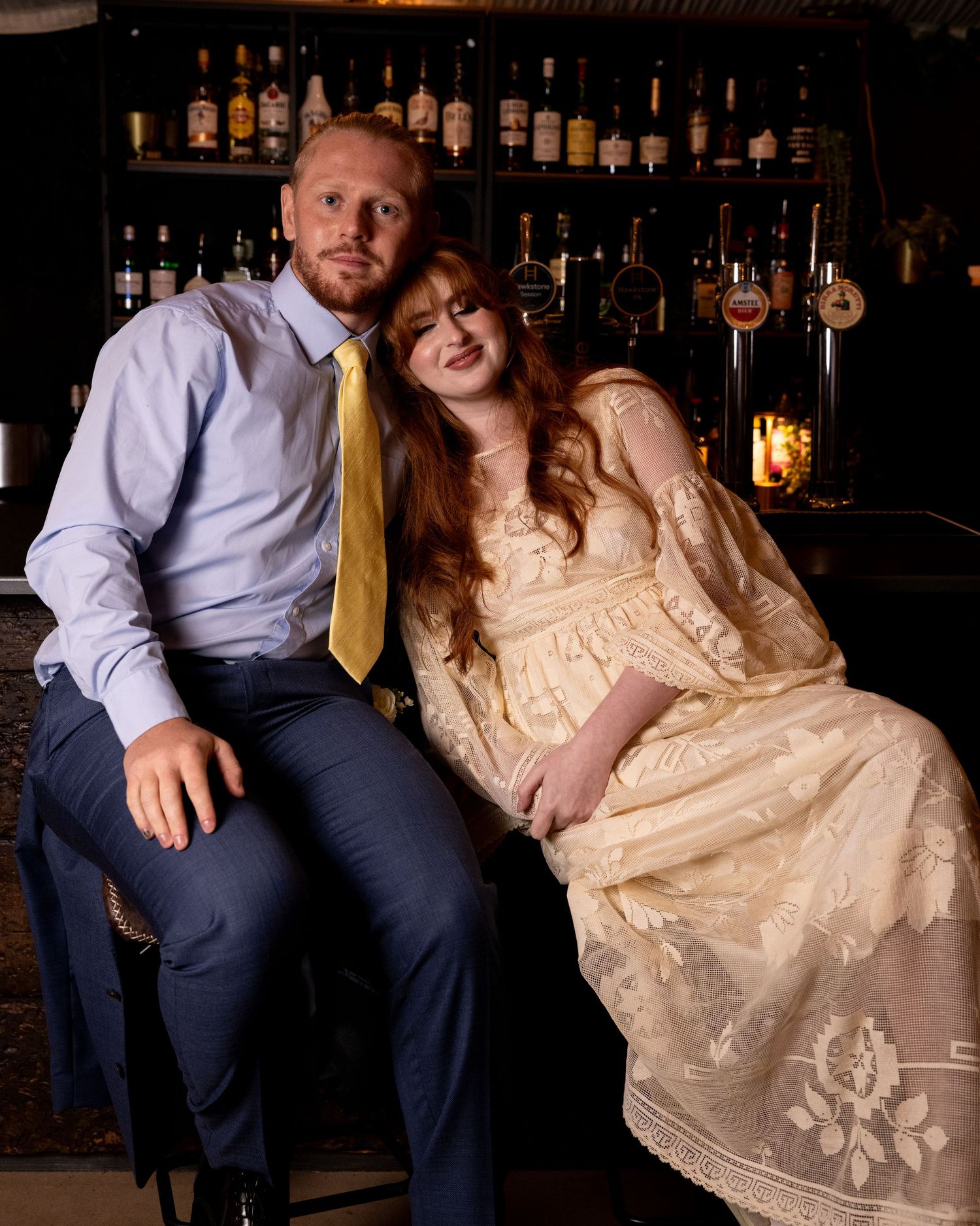 Couple in formal attire sitting together at a bar, woman in cream vintage dress leaning on man.