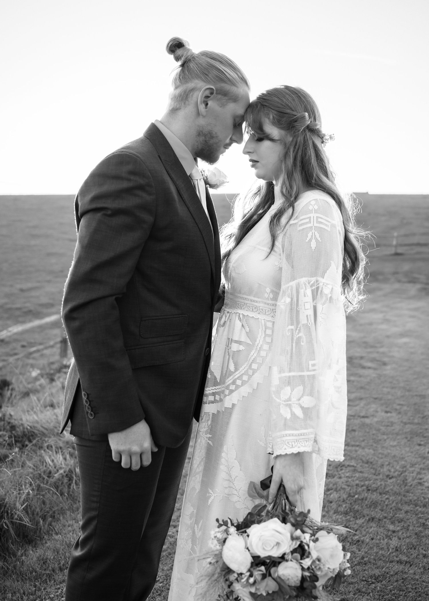 Black and white photo of bride and groom facing each other near the ocean holding a bouquet.