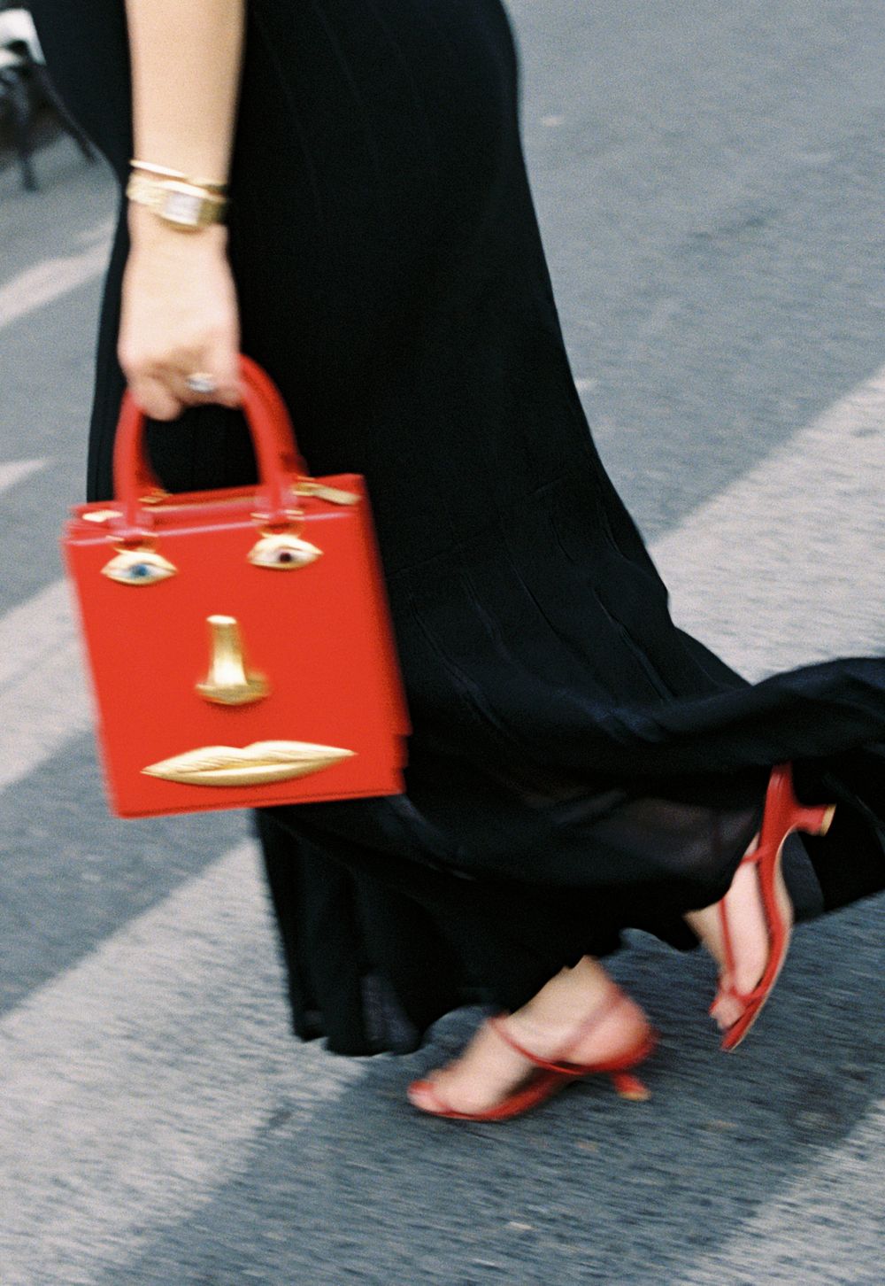 Woman in black dress carrying a red Schiaparelli  face handbag with gold details, paired with red heeled sandals. Paris outfit ideas, film