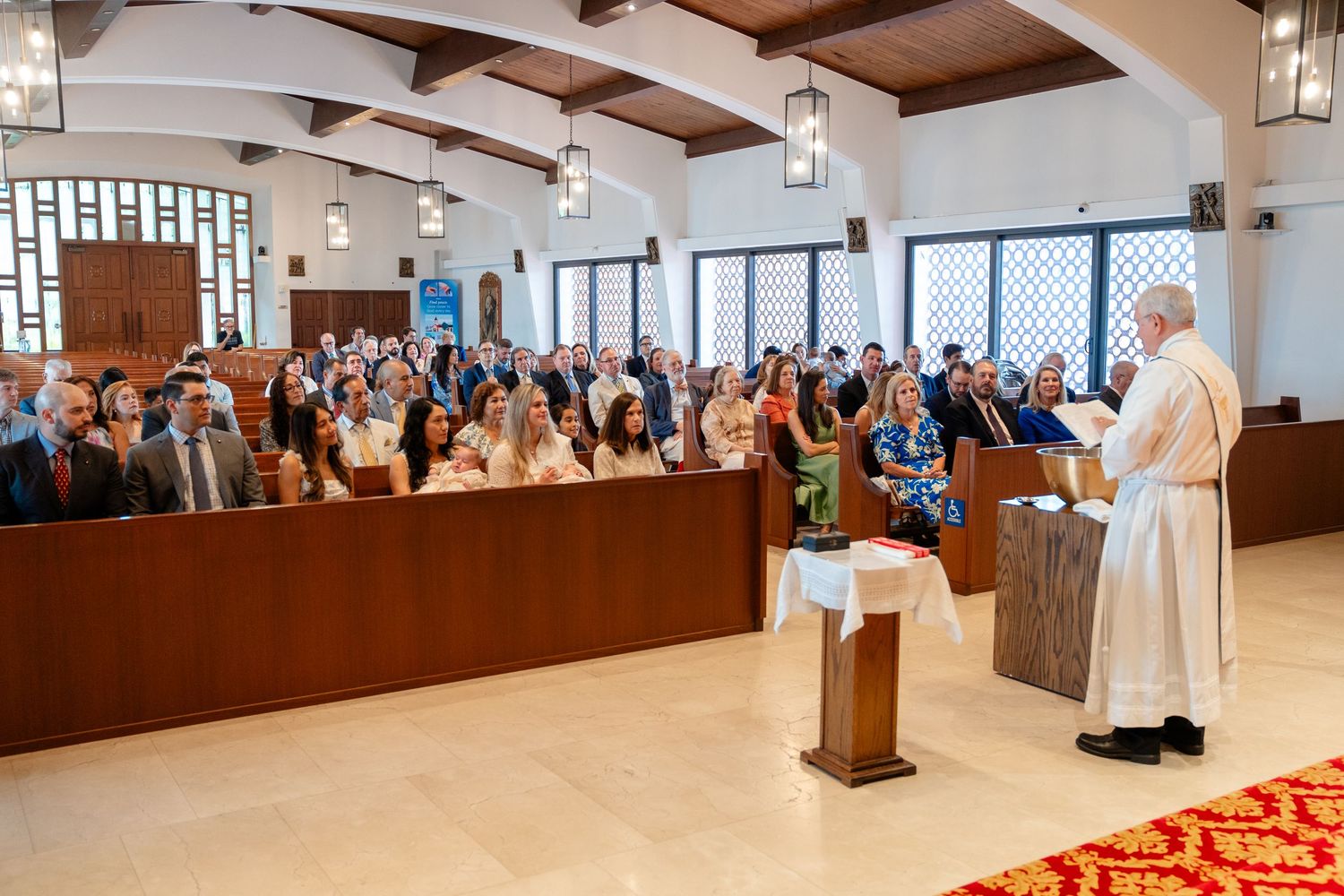 Priest addresses congregation during Catholic Mass ceremony in modern church with wooden pews filled with attendees.