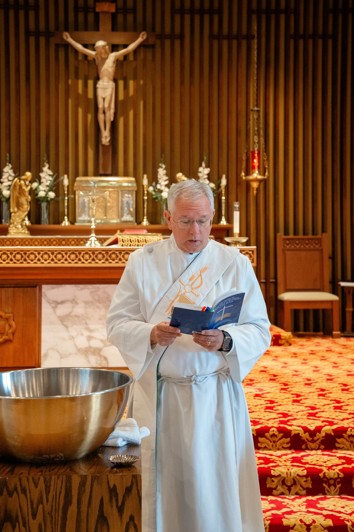 Priest in white vestments reads from a blue book at a baptismal font inside a Catholic church altar.