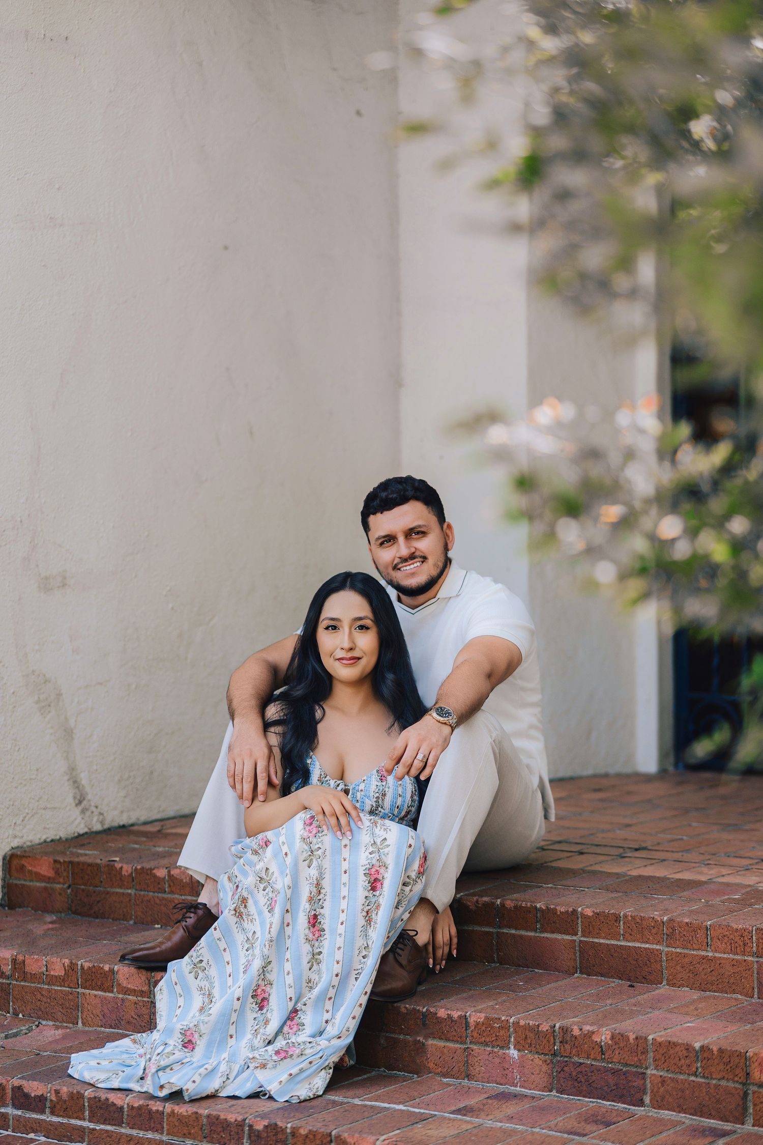 A couple sits together on brick steps, the woman in a floral blue dress, the man in white, smiling warmly at the camera.