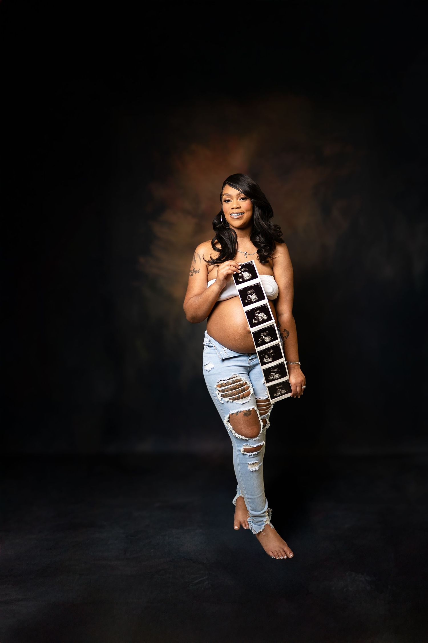 Pregnant woman in ripped jeans holding ultrasound photos during a professional maternity photo shoot on dark background.
