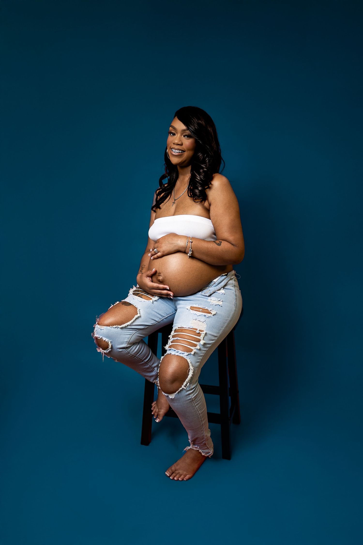 Pregnant woman in white bandeau top and ripped jeans cradling baby bump, seated on stool against teal background.