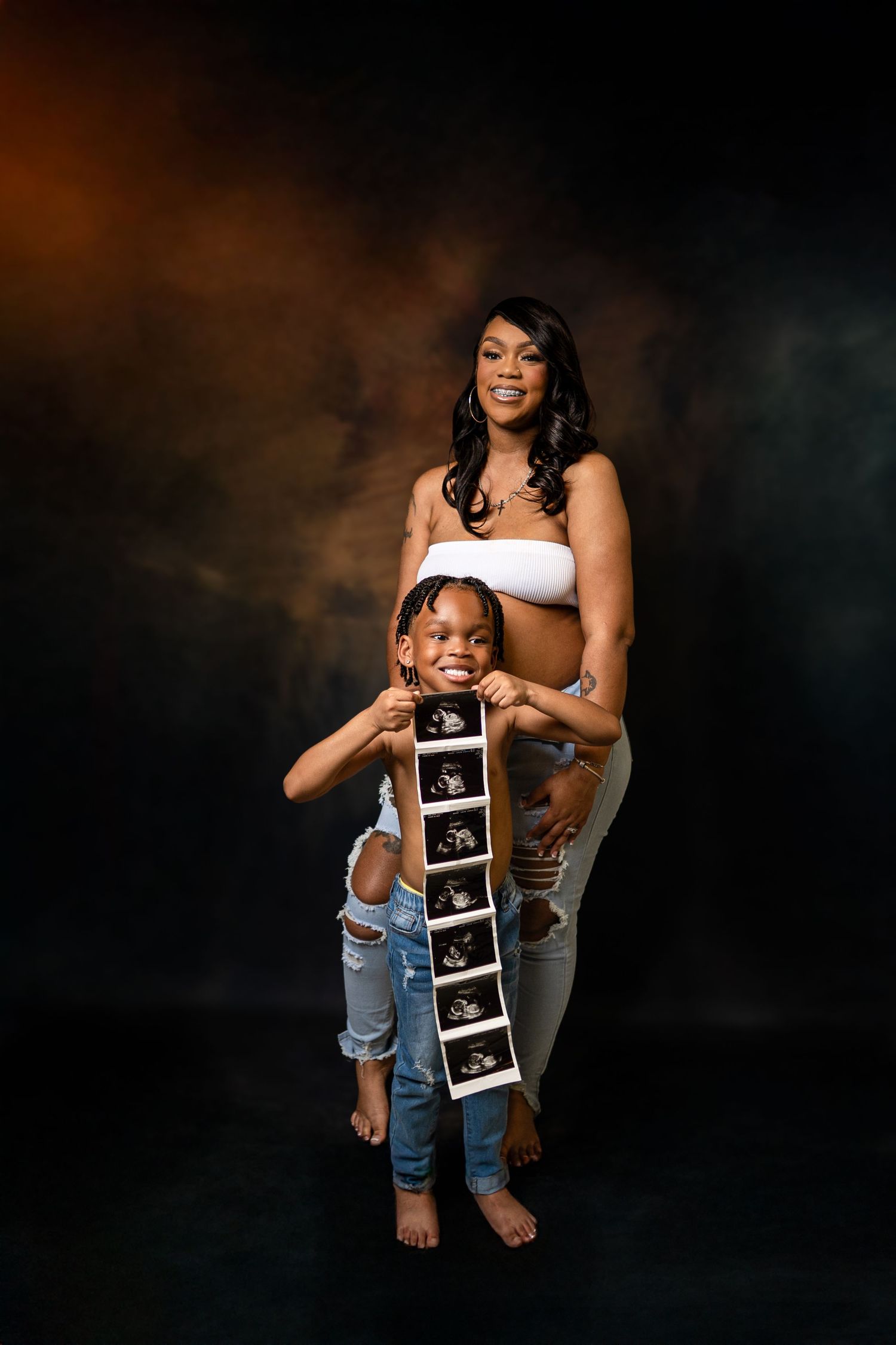 Pregnant woman in white bandeau and ripped jeans poses with young girl holding ultrasound strip in studio.