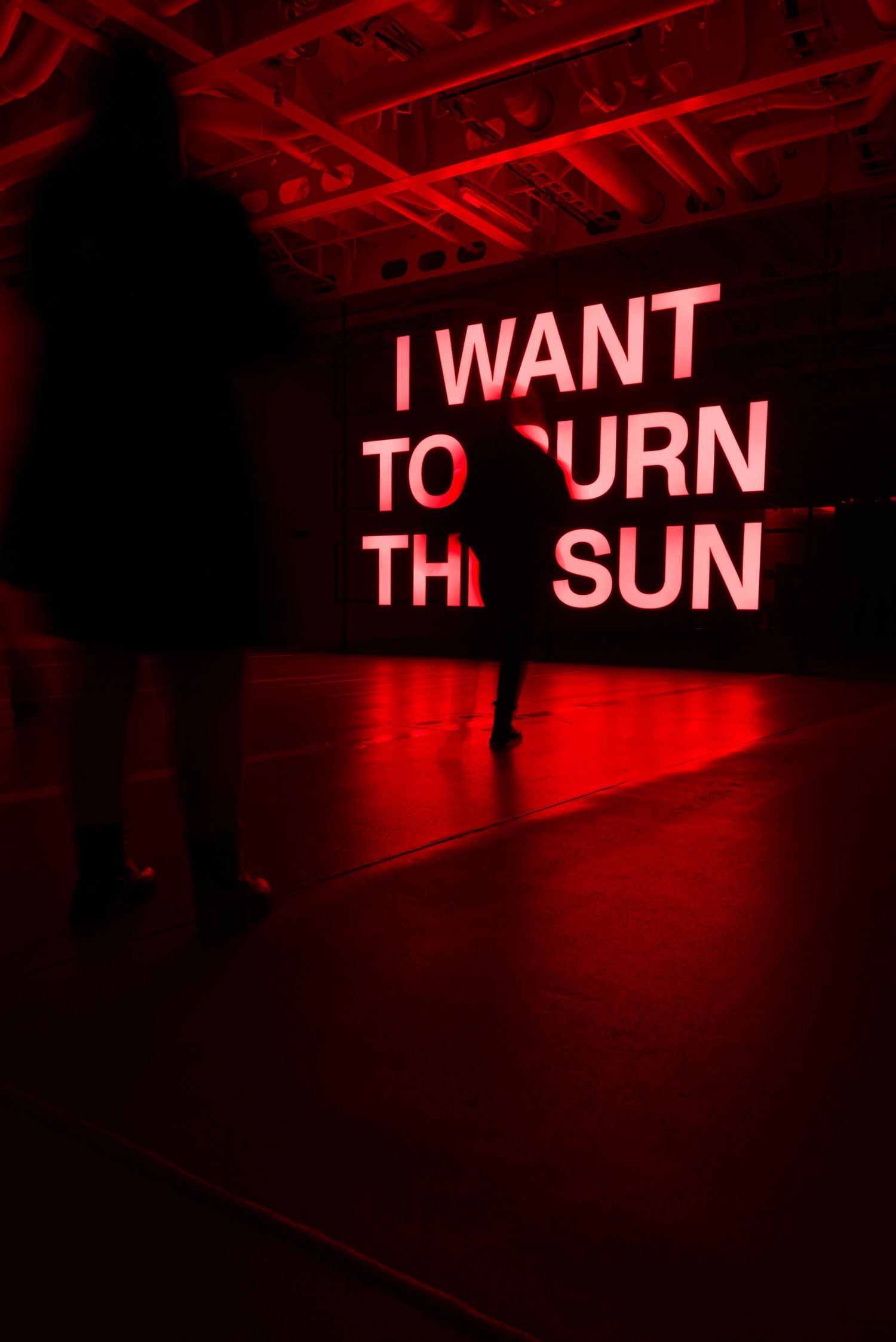 Red neon text installation reading I Want To Burn The Sun with silhouetted figures in a darkened gallery space.