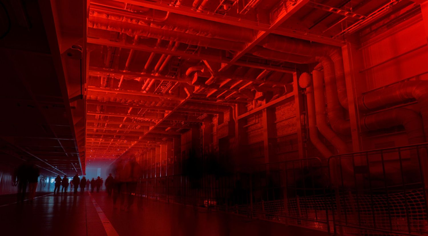 Dramatic red-lit industrial hall with silhouetted figures and glowing circuit-board patterns on walls and ceiling.