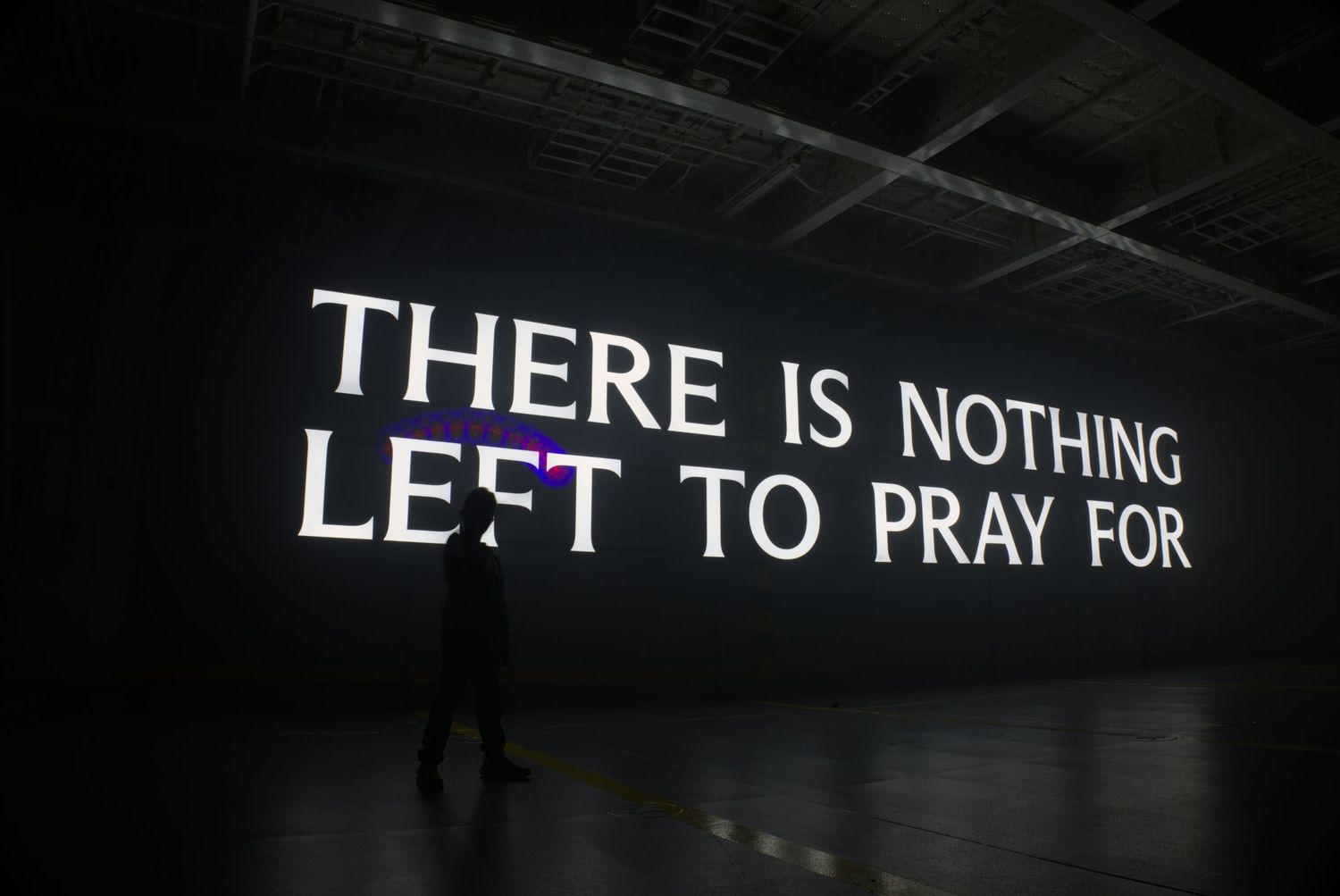 Dark gallery space with illuminated text projection reading THERE IS NOTHING LEFT TO PRAY FOR with silhouetted figure.