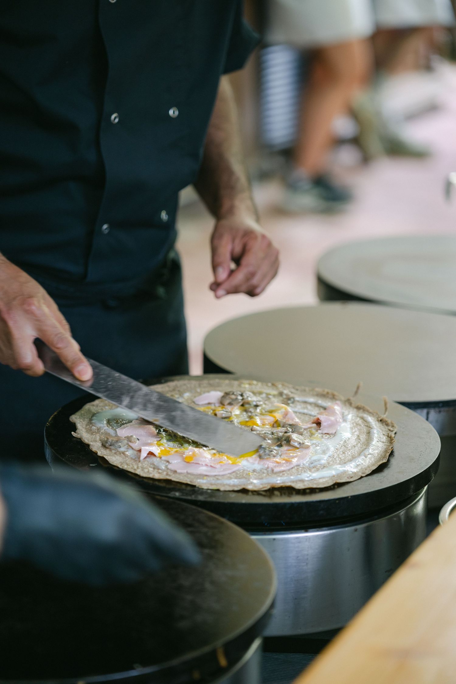 Chef spreading batter on a hot crepe griddle, preparing a savory crepe with colorful toppings at a food stall.