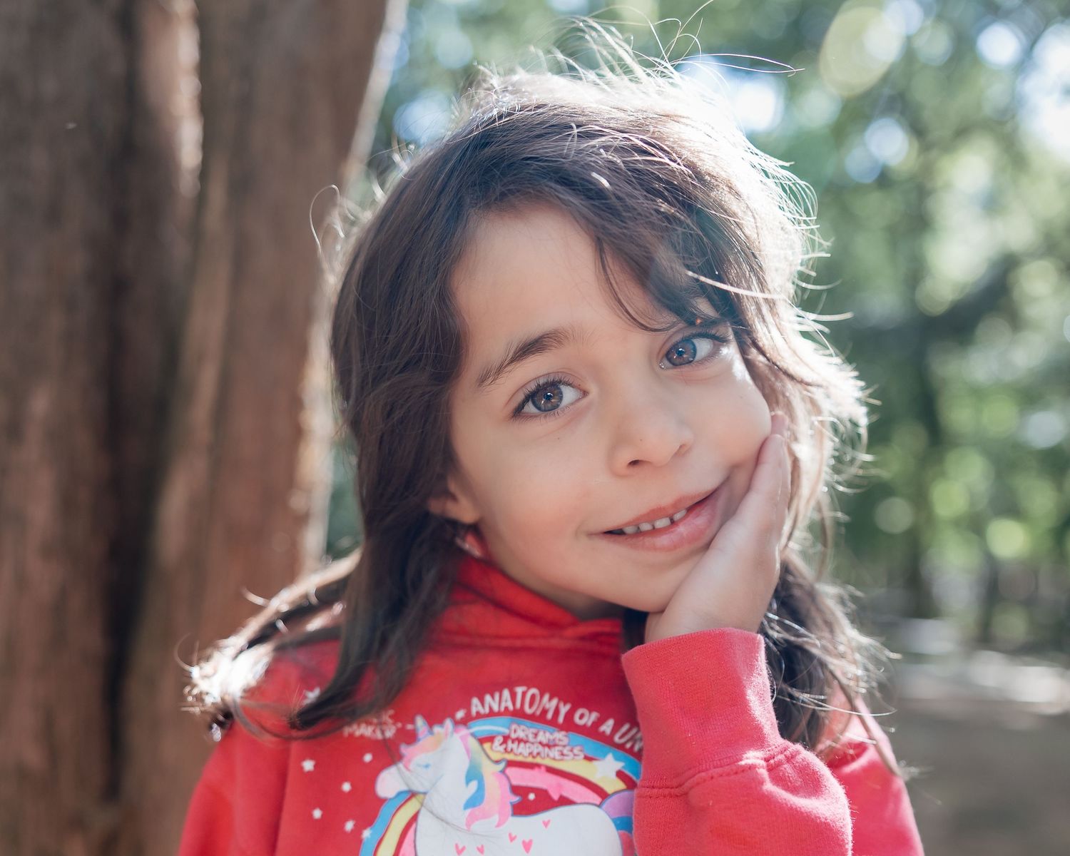 A smiling young girl with brown hair wearing a red unicorn hoodie poses outdoors near trees.