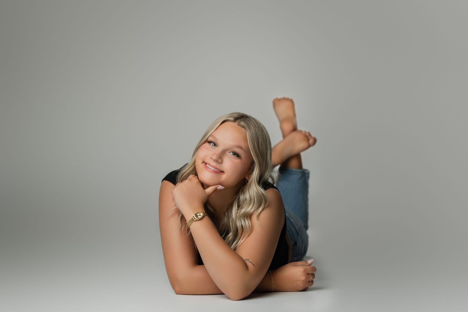 Smiling blonde girl lying on floor in jeans, resting chin on hands, studio portrait with gray background.
